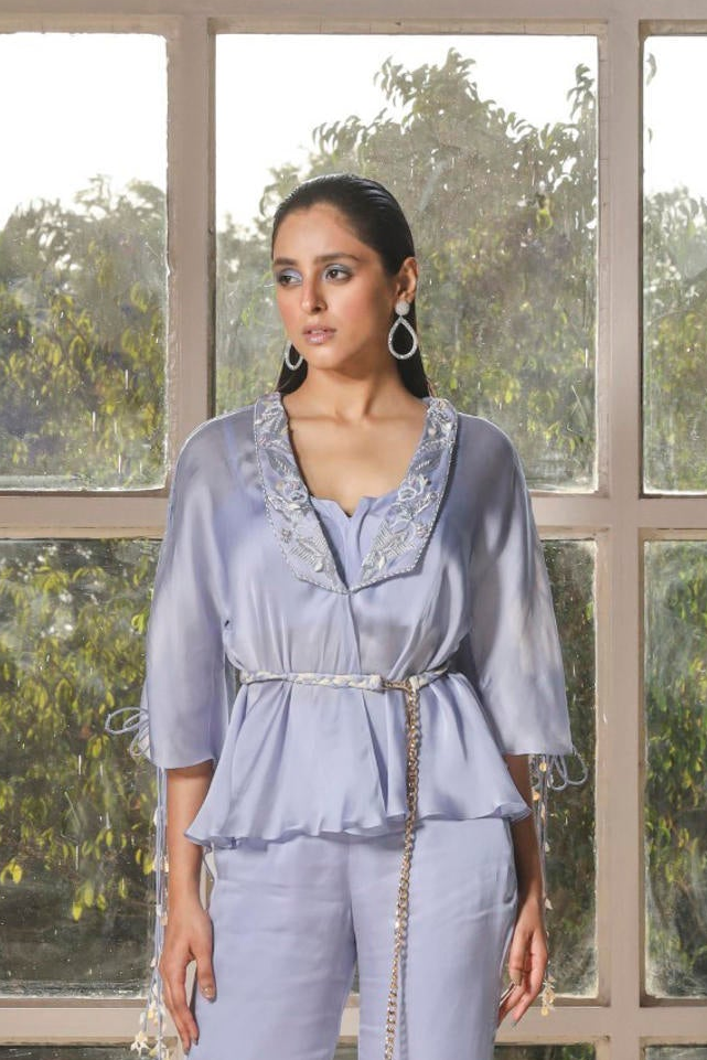 A woman wearing a periwinkle blue co-ord set with embroidery, consisting of a kimono top, pants, and a hand braided belt, standing in front of a large window.