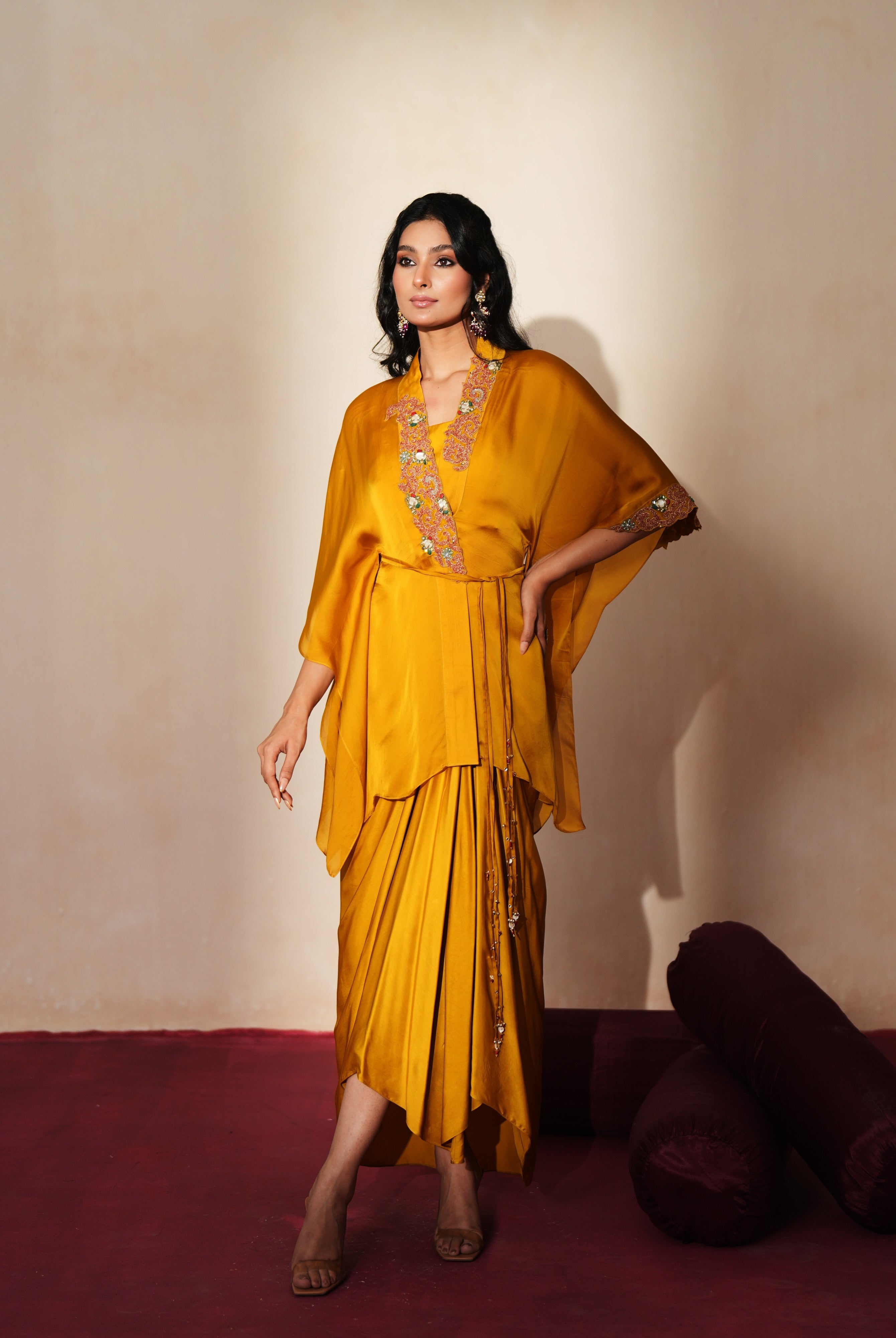 Woman in a golden yellow traditional outfit standing againsta beige wall on a red carpet.