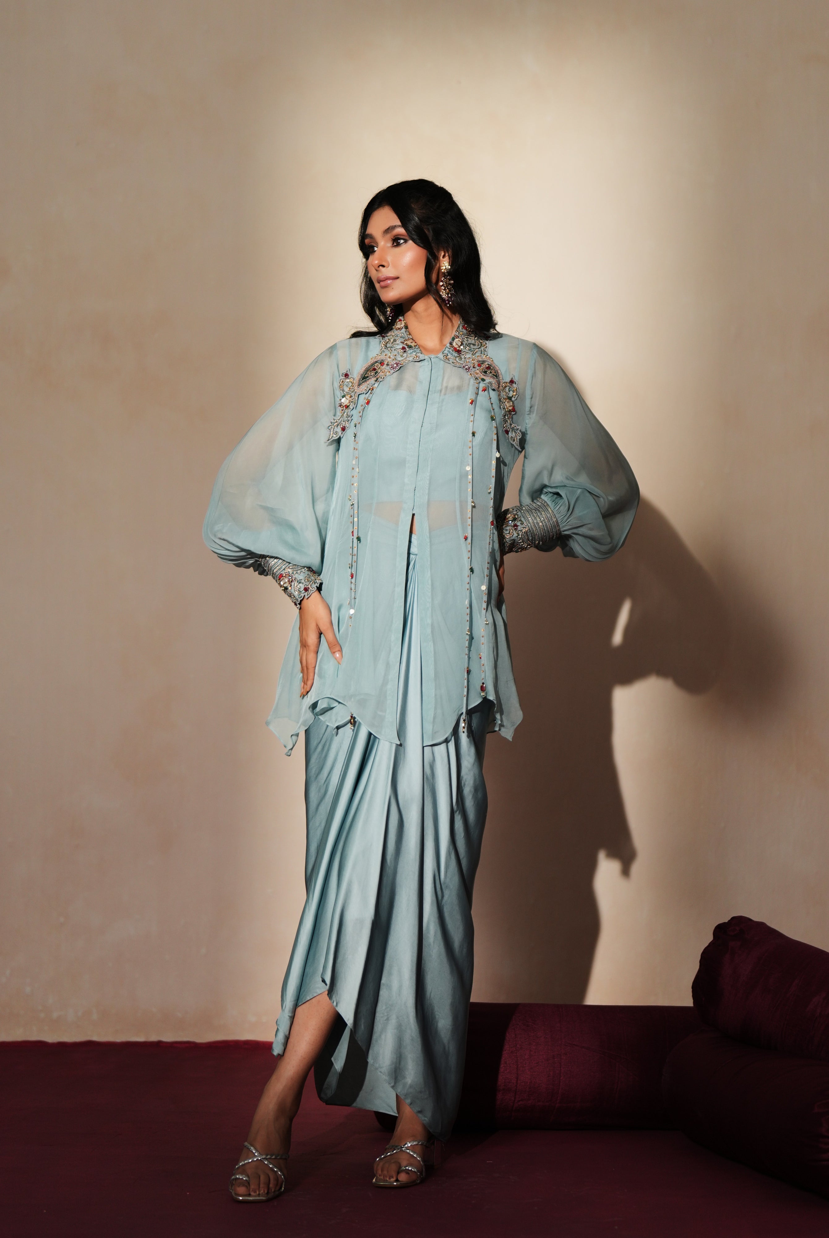 Woman in a pastel blue traditional outfit standing againsta beige wall on a red carpet.