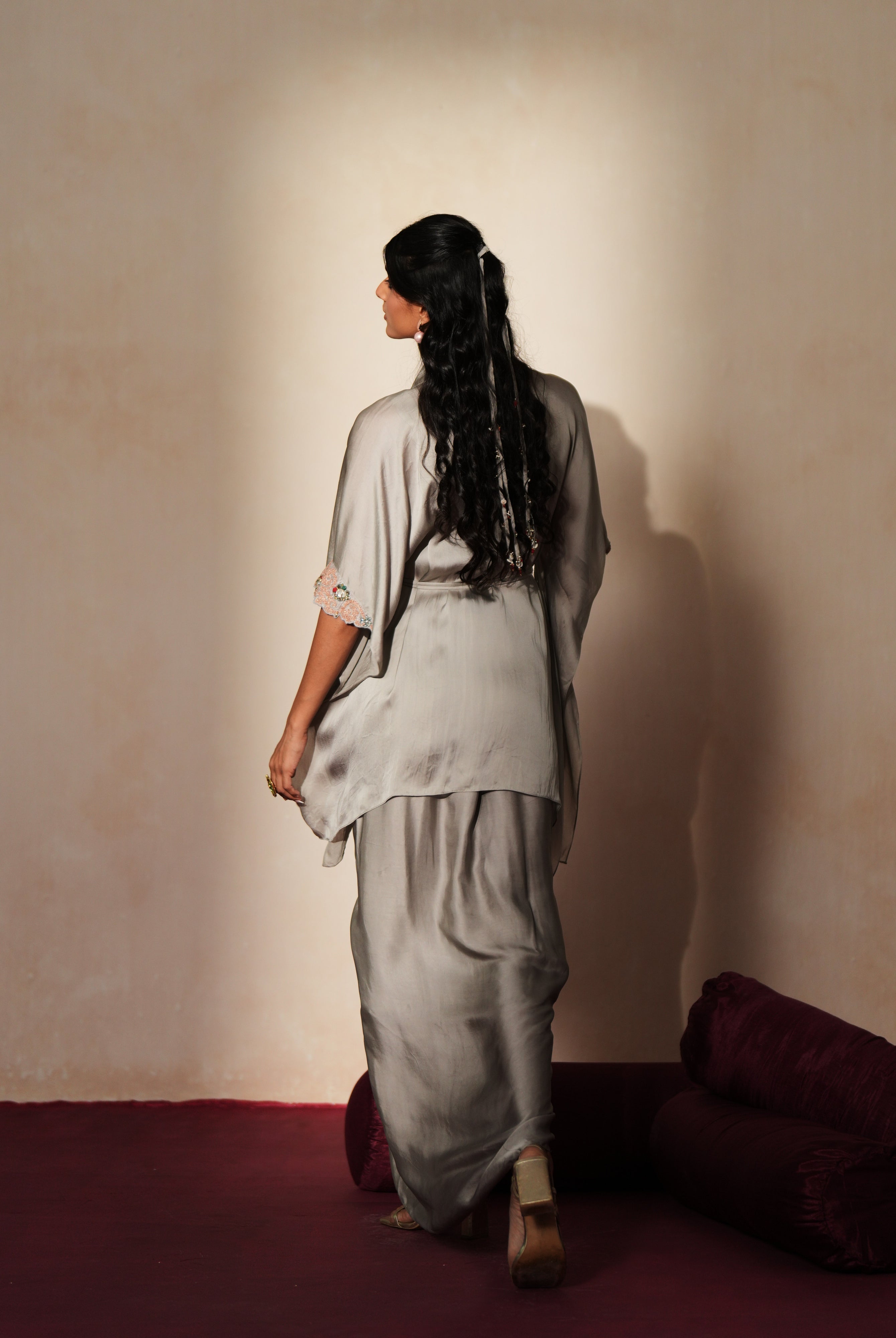 Woman in a mettalic grey traditional outfit standing againsta beige wall on a red carpet.