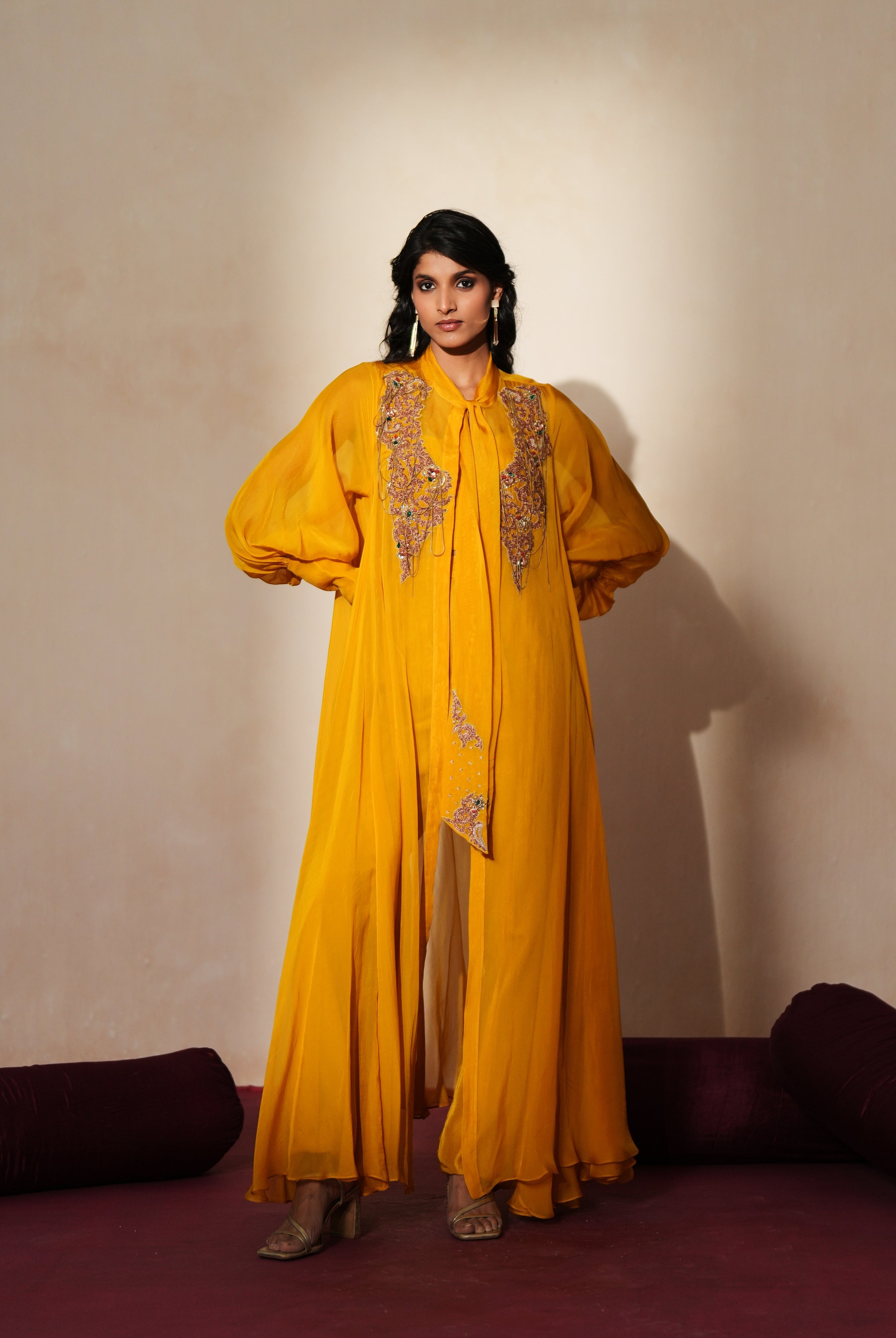 Woman in a golden yellow traditional outfit standing againsta beige wall on a red carpet.