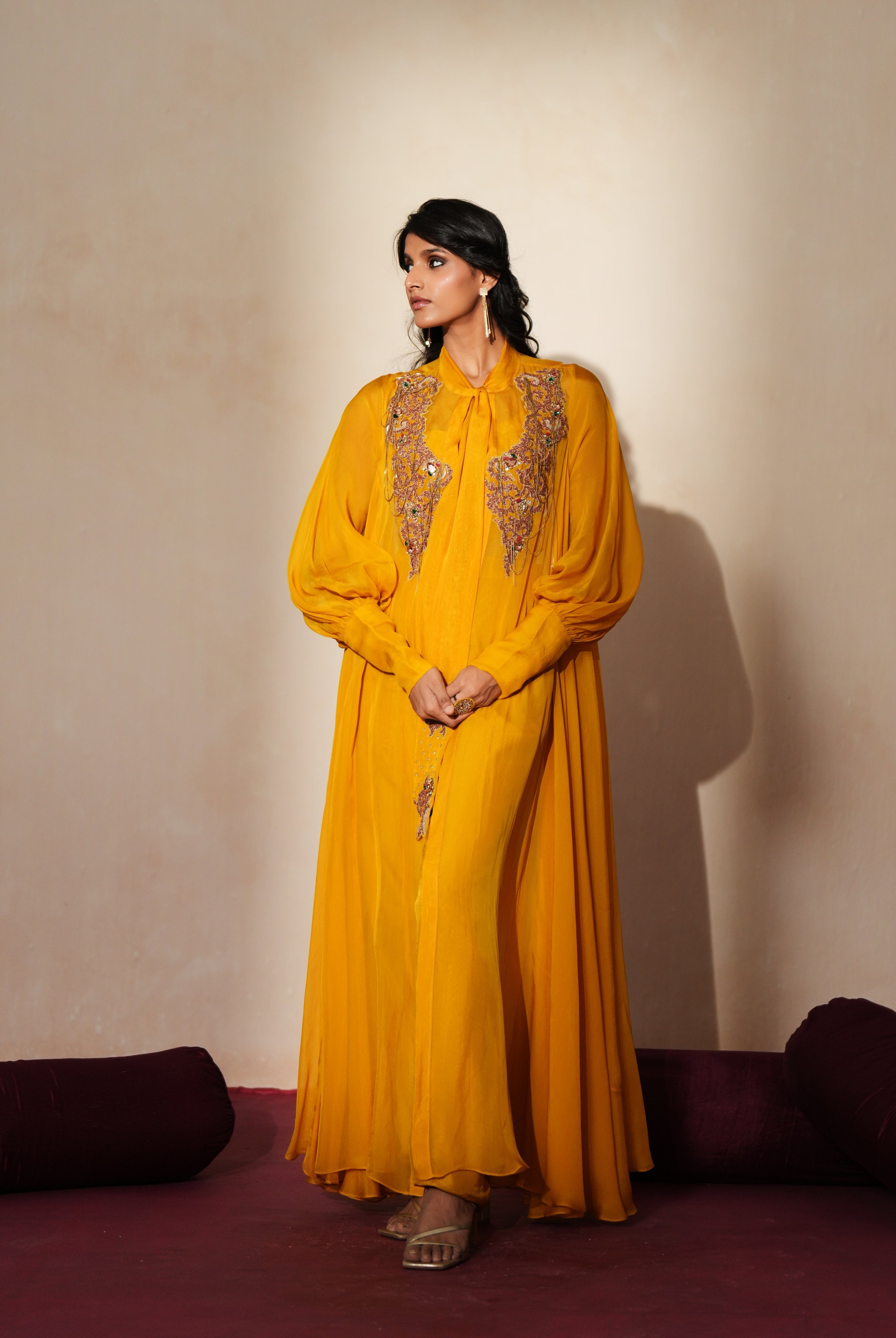 Woman in a golden yellow traditional outfit standing againsta beige wall on a red carpet.