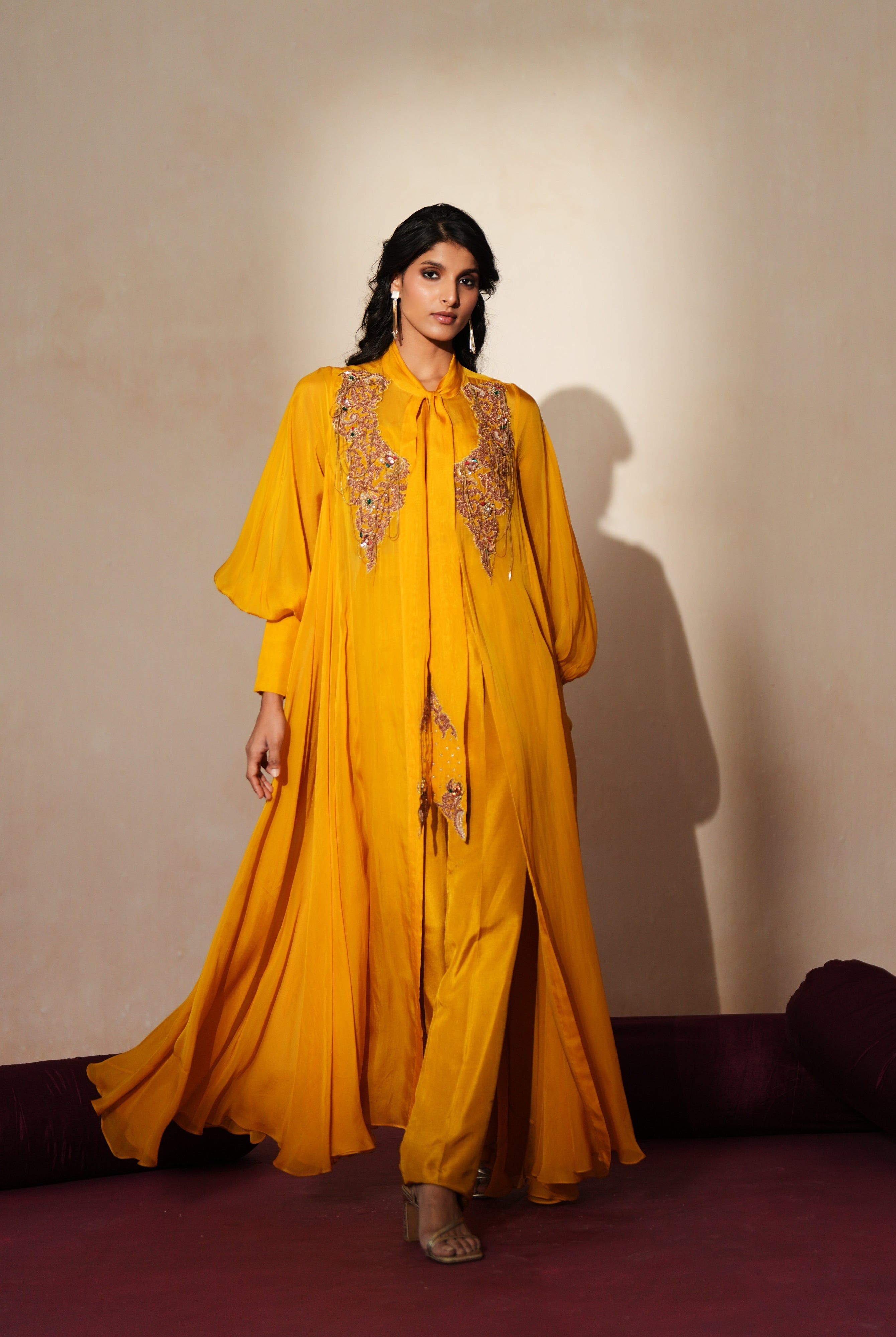 Woman in a golden yellow traditional outfit standing againsta beige wall on a red carpet.