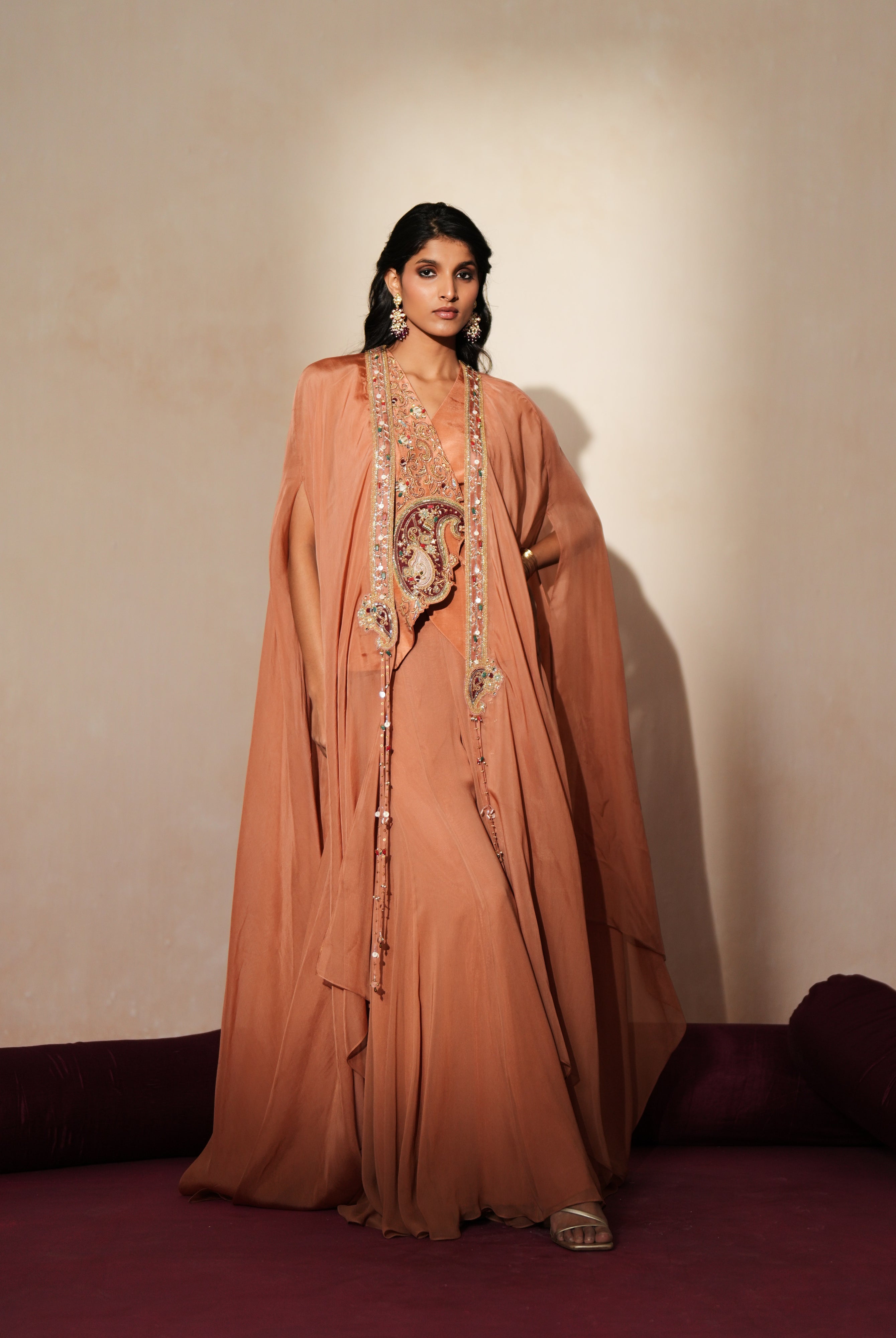 Woman in a metallic mocha traditional outfit standing againsta beige wall on a red carpet.