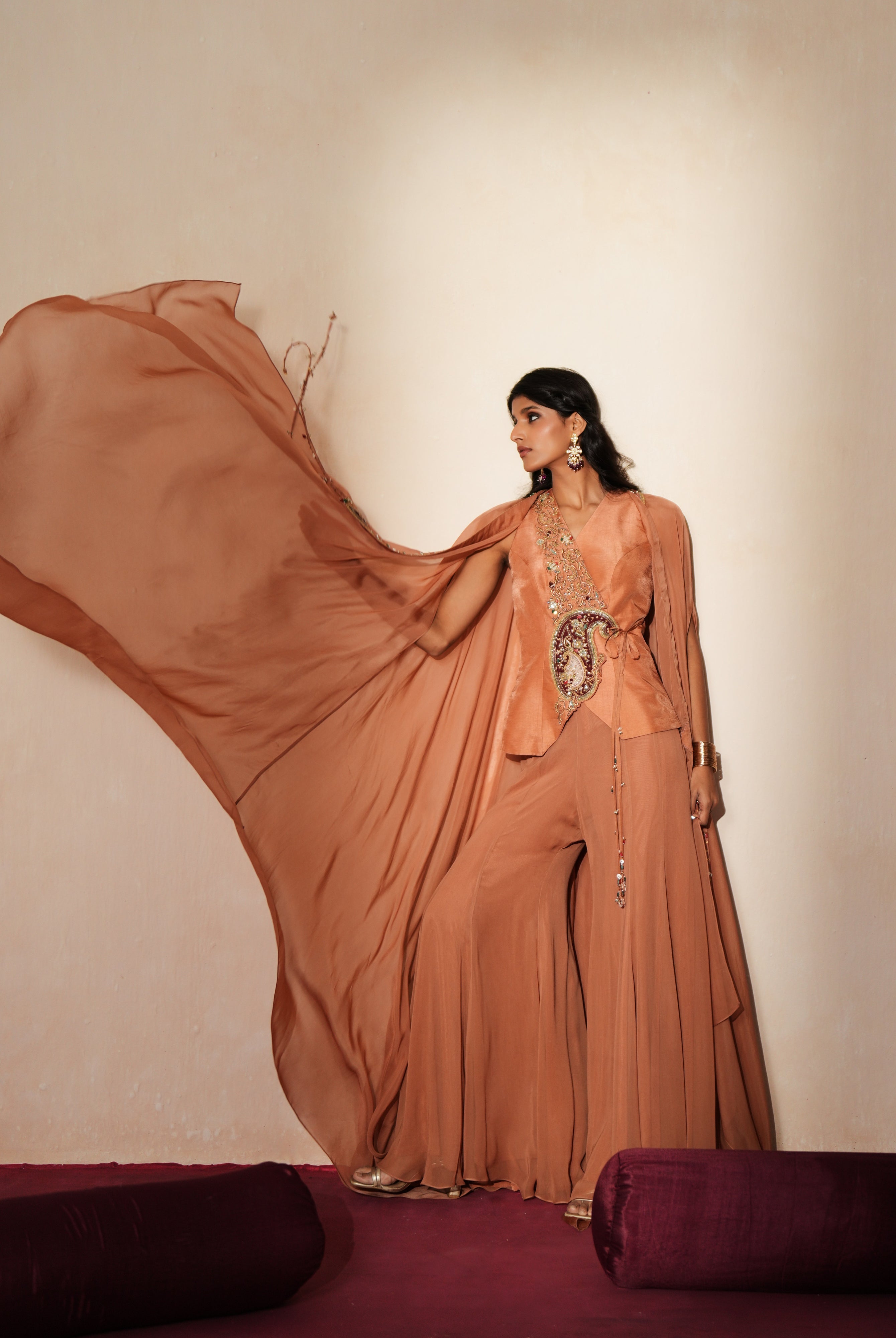 Woman in a metallic mocha traditional outfit standing againsta beige wall on a red carpet.