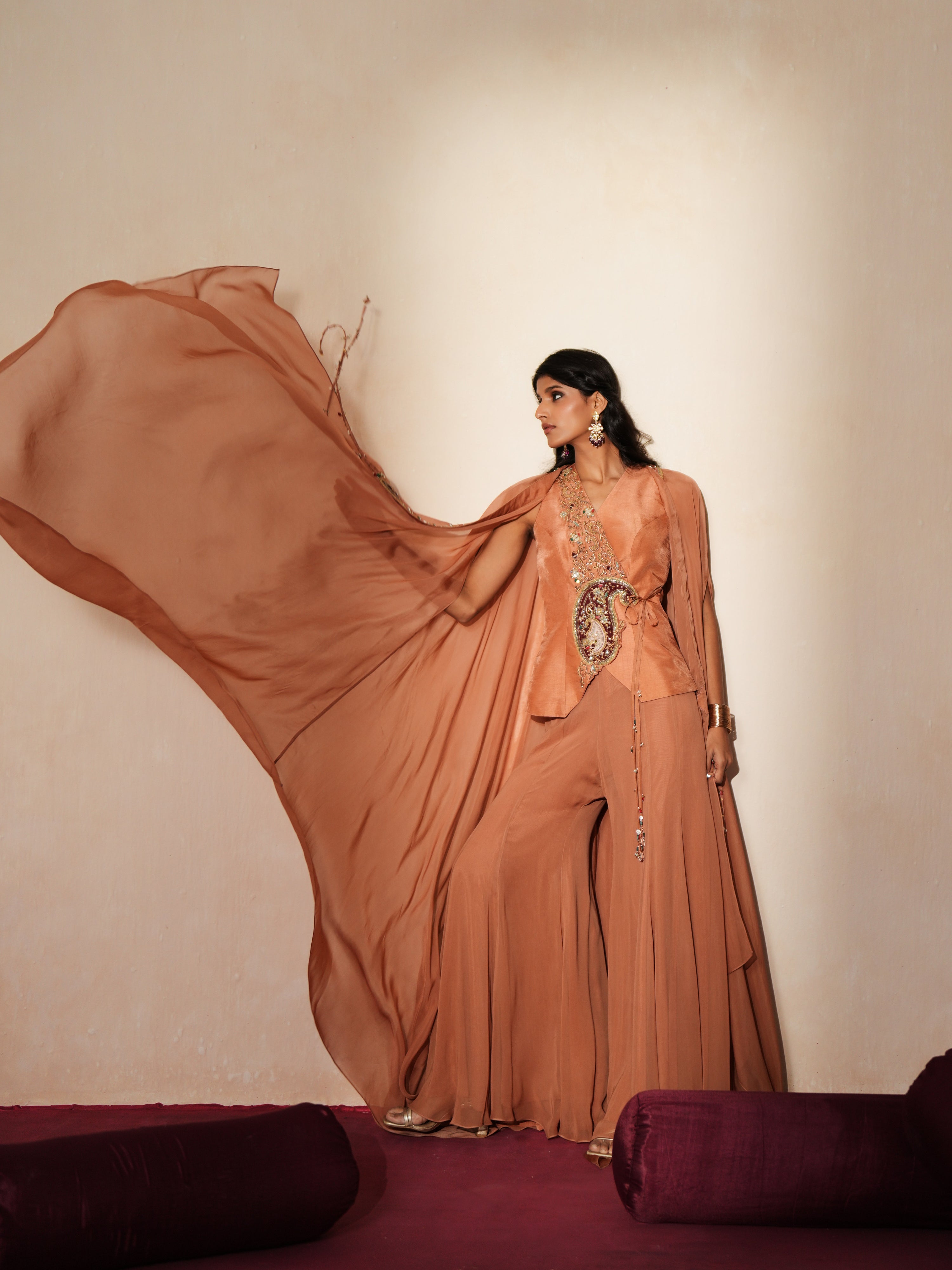 Woman in a metallic mocha traditional outfit standing againsta beige wall on a red carpet.
