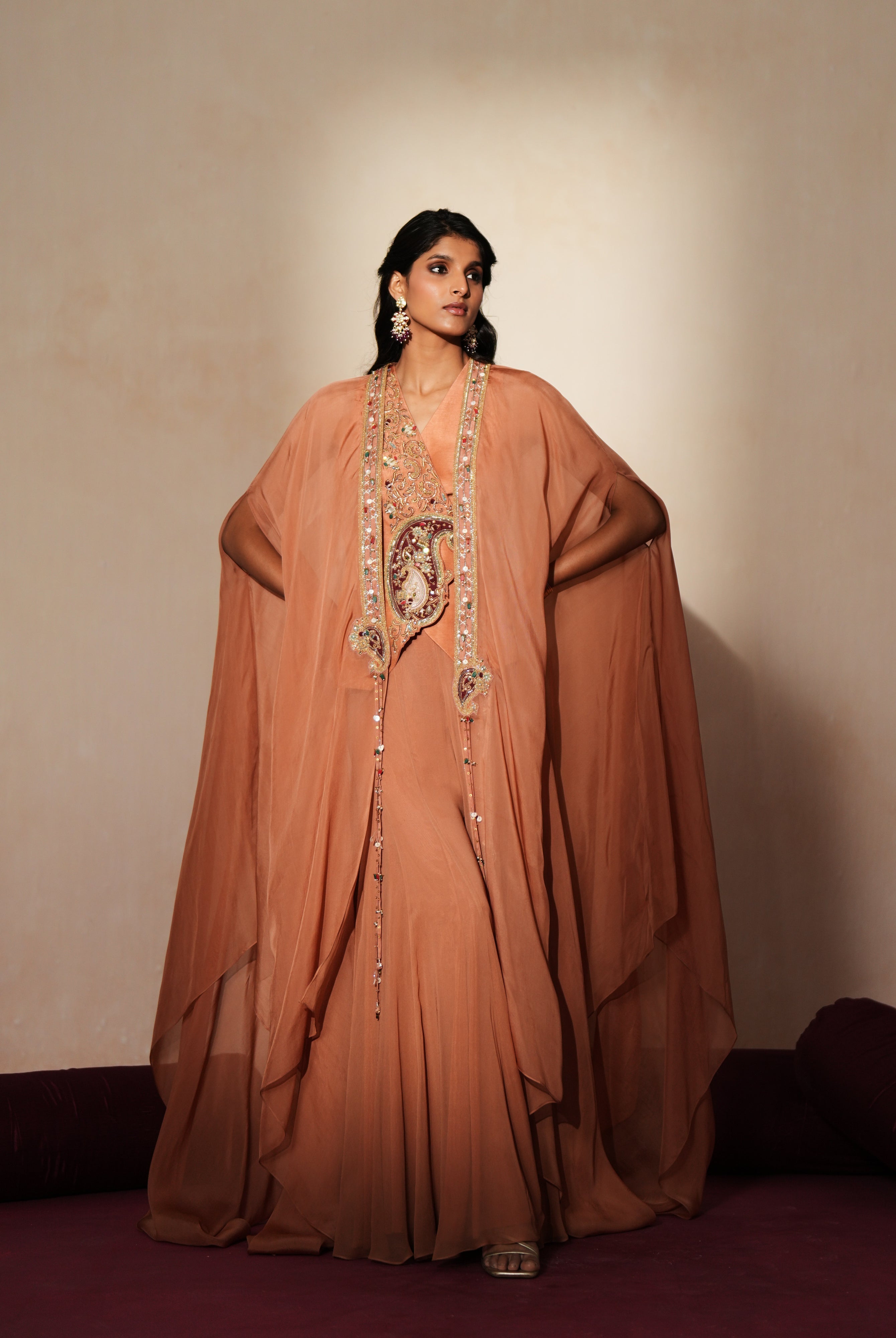 Woman in a metallic mocha traditional outfit standing againsta beige wall on a red carpet.