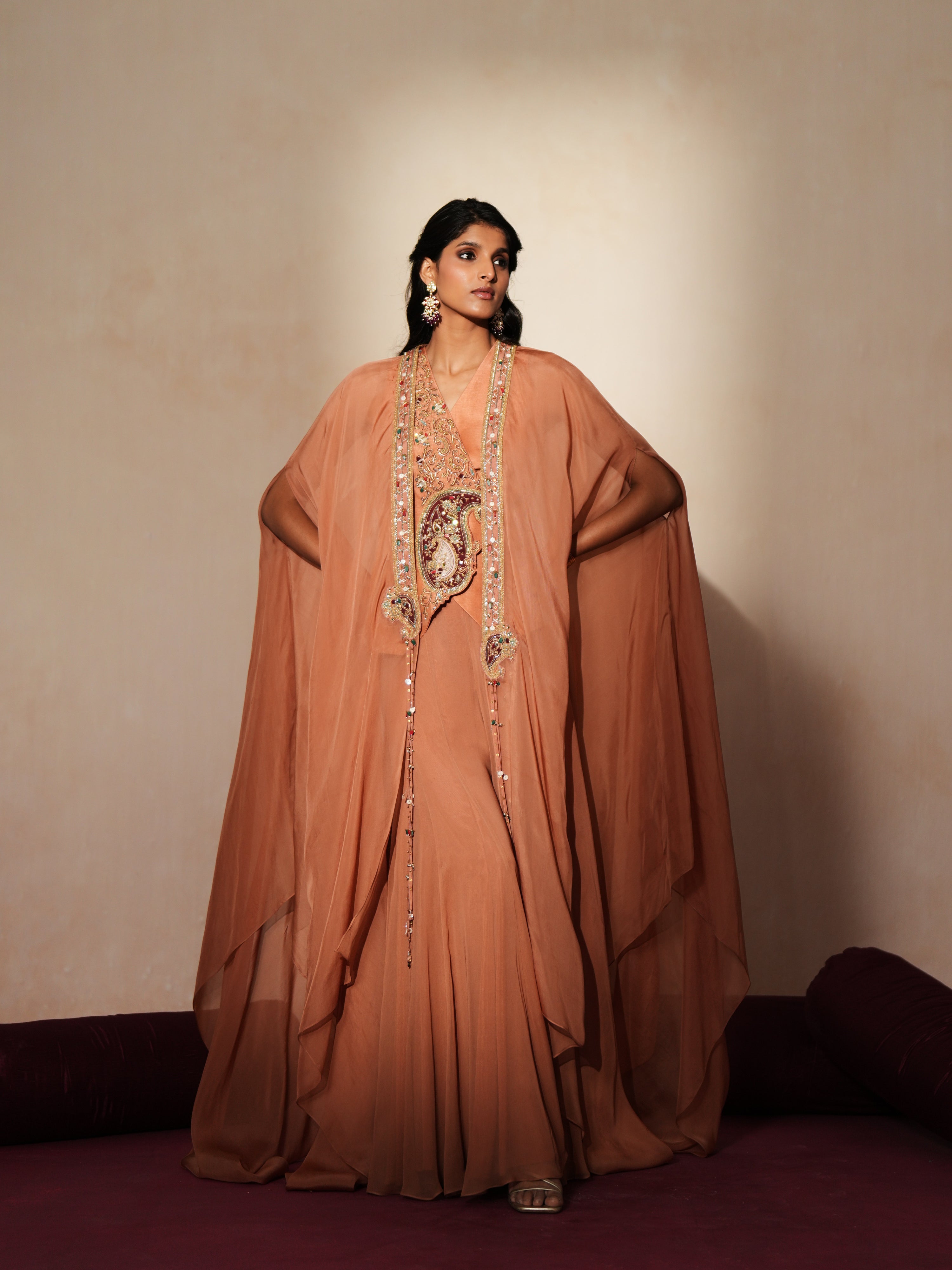 Woman in a metallic mocha traditional outfit standing againsta beige wall on a red carpet.
