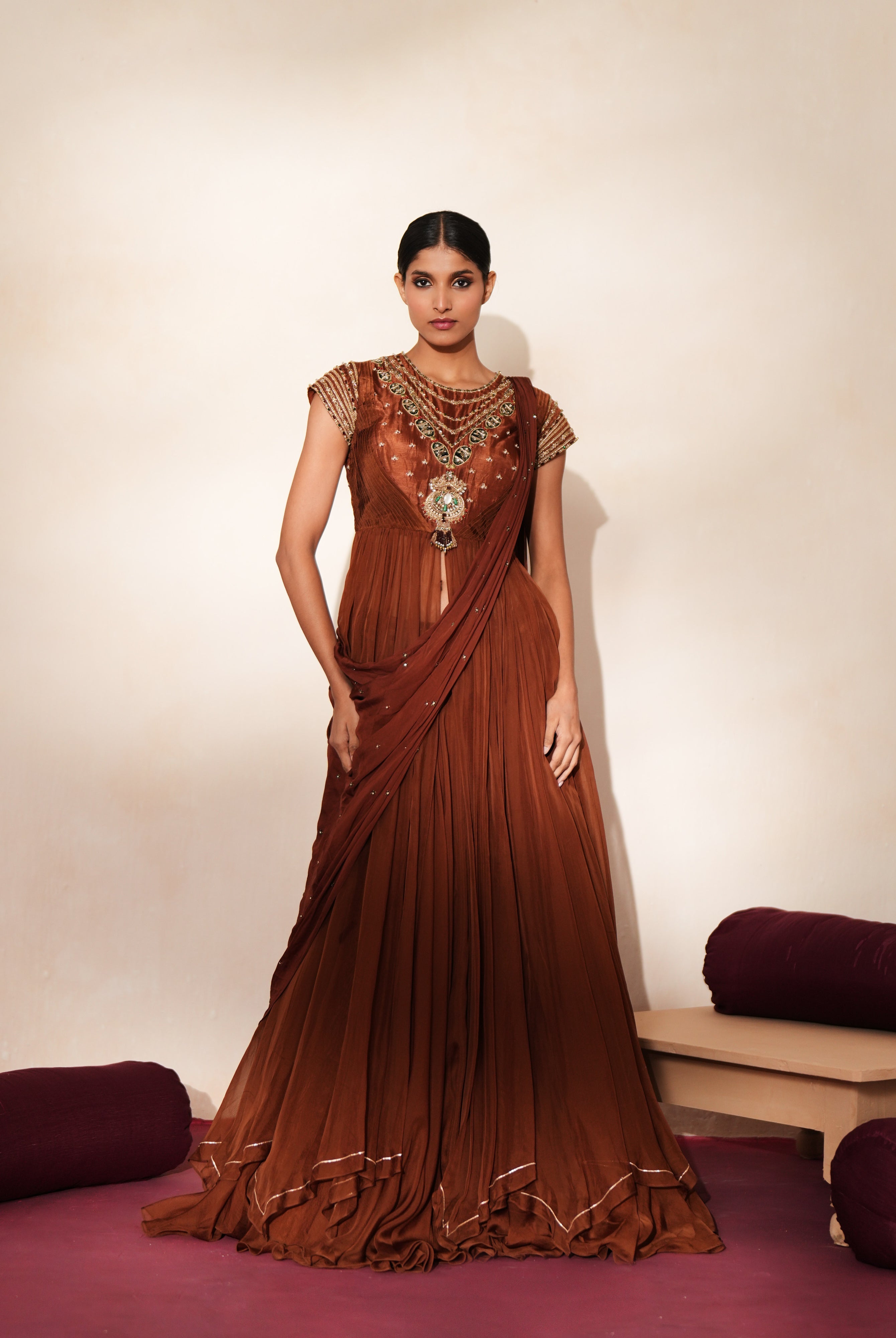 Woman in a mettalic brown traditional outfit standing againsta beige wall on a red carpet.