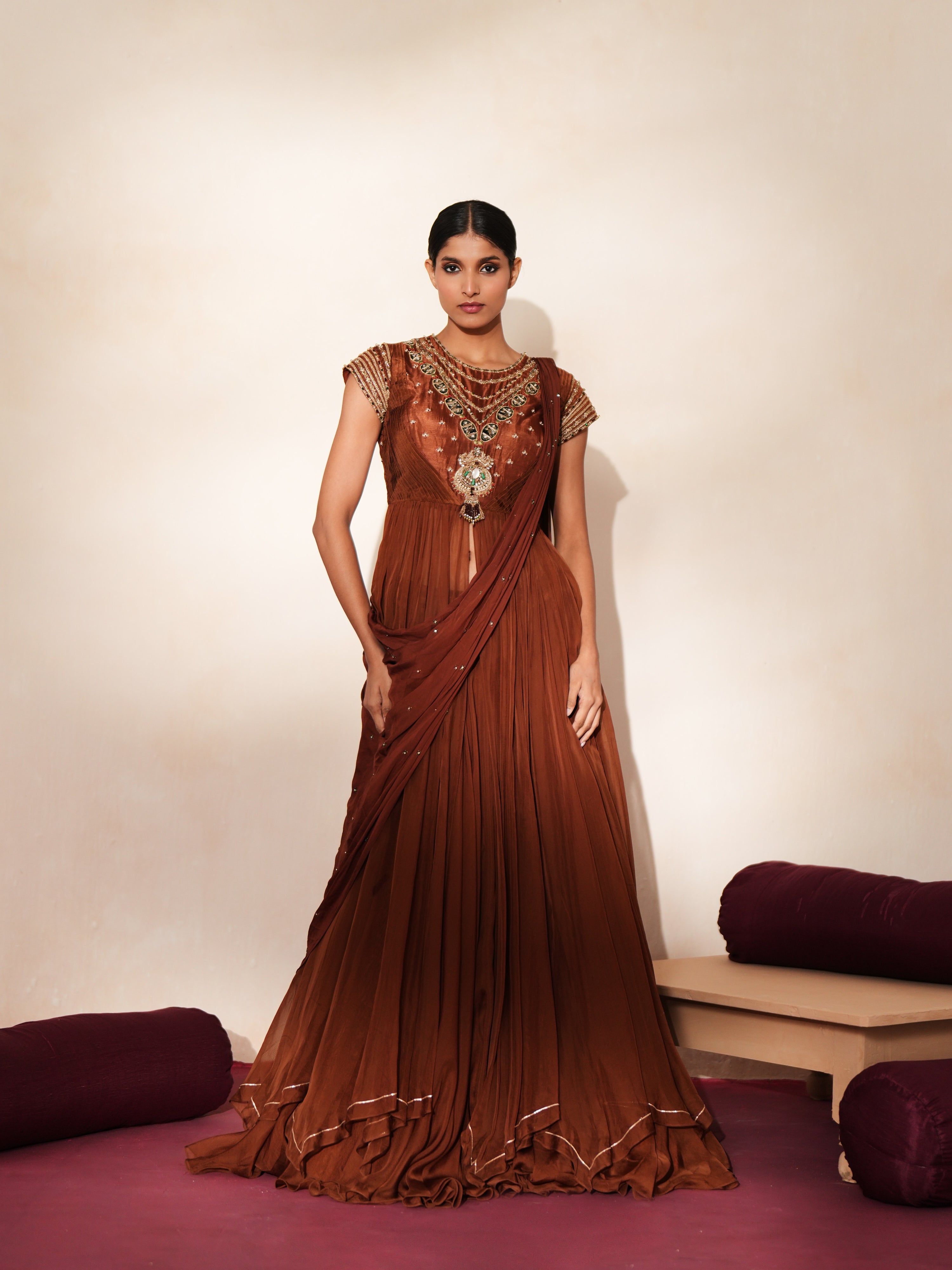 Woman in a mettalic brown traditional outfit standing againsta beige wall on a red carpet.