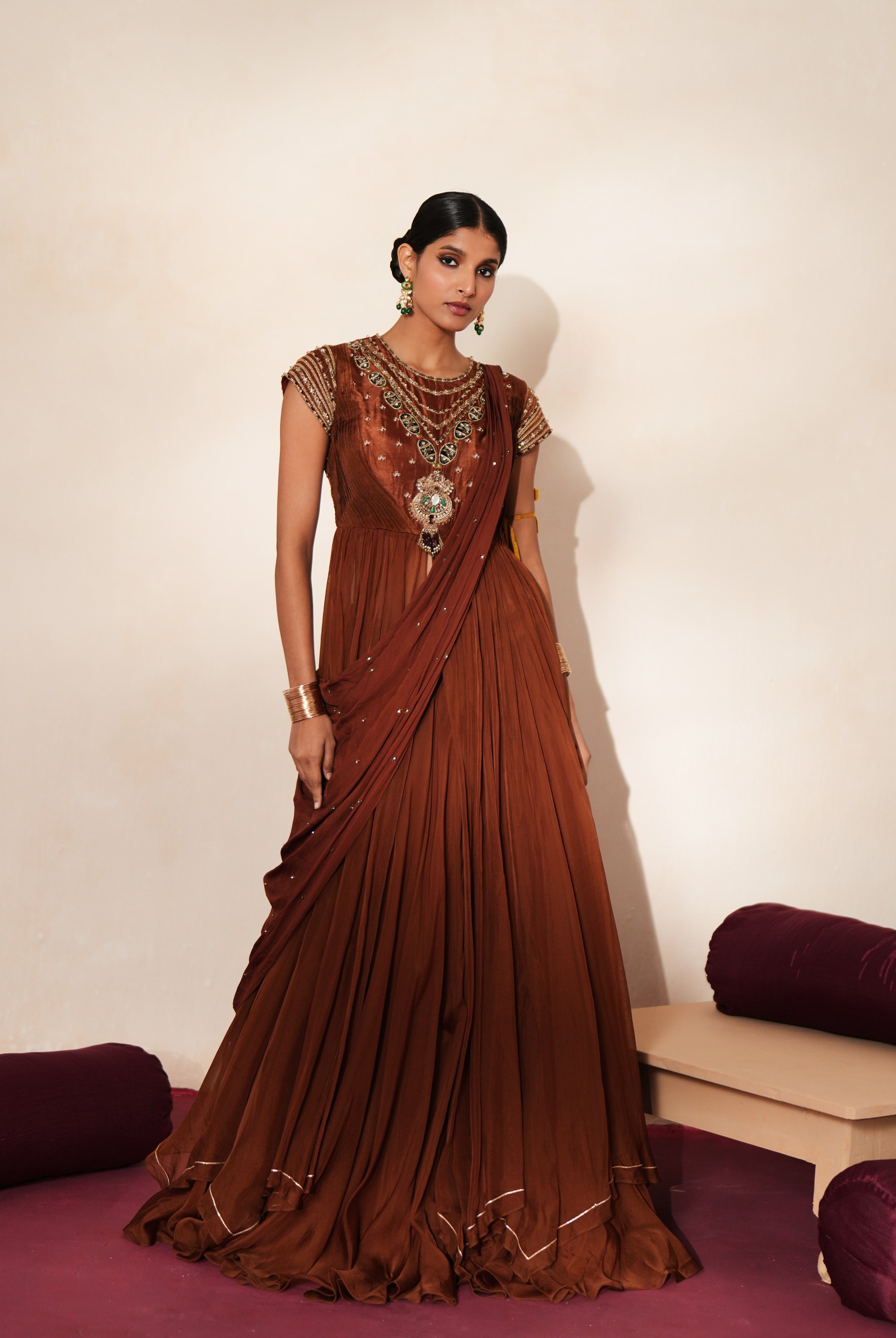 Woman in a mettalic brown traditional outfit standing againsta beige wall on a red carpet.
