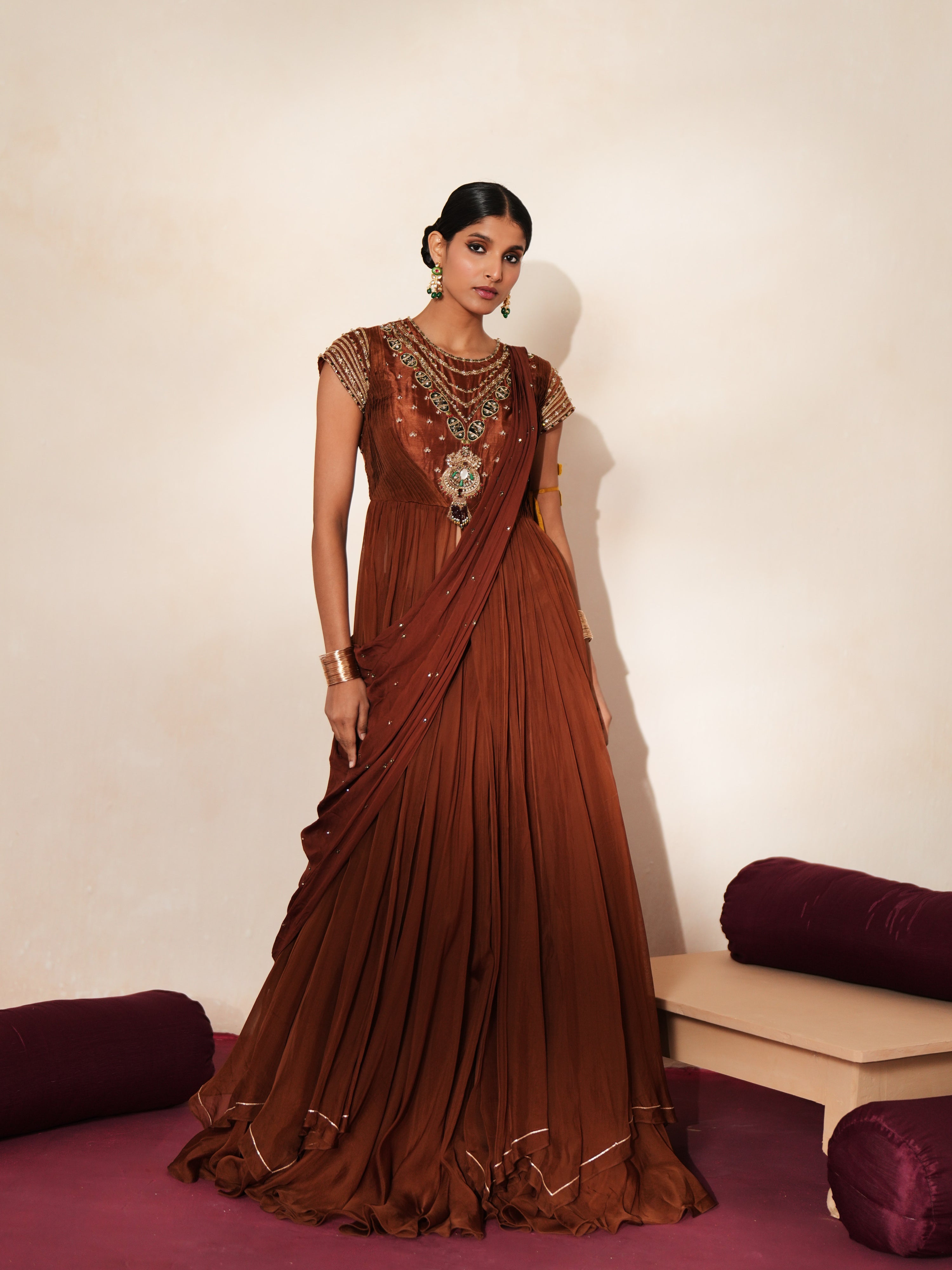 Woman in a mettalic brown traditional outfit standing againsta beige wall on a red carpet.