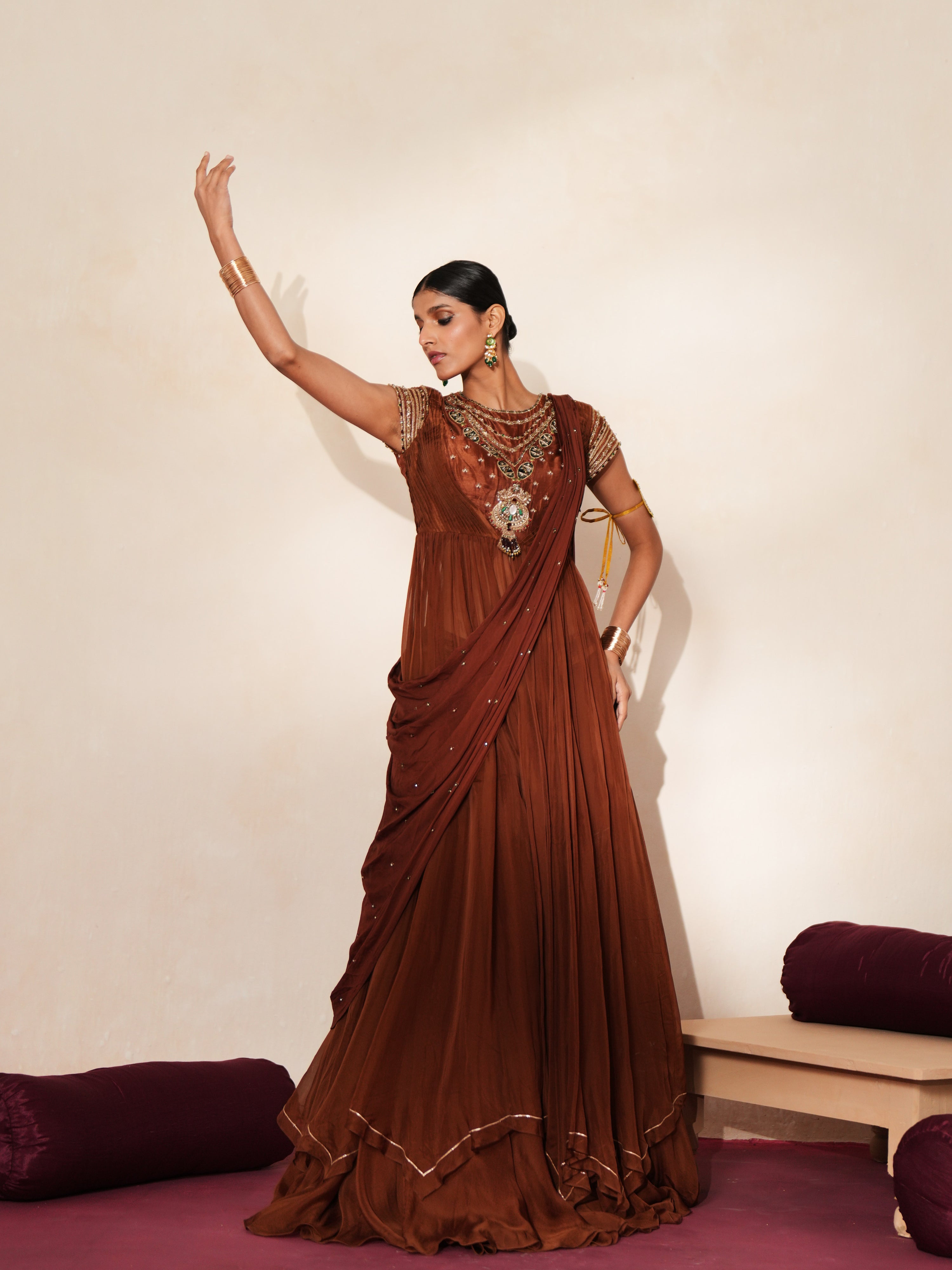 Woman in a mettalic brown traditional outfit standing againsta beige wall on a red carpet.