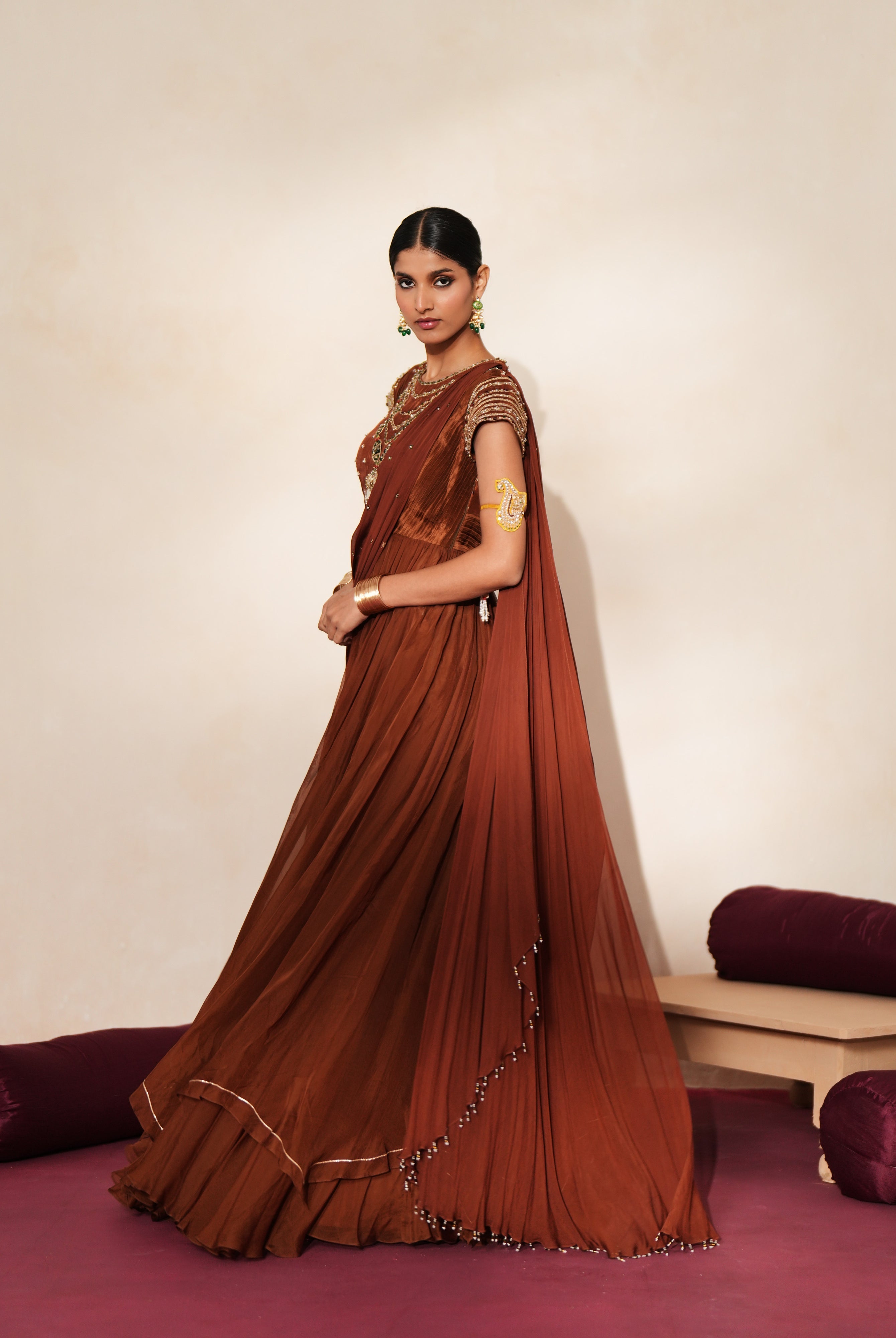 Woman in a mettalic brown traditional outfit standing againsta beige wall on a red carpet.