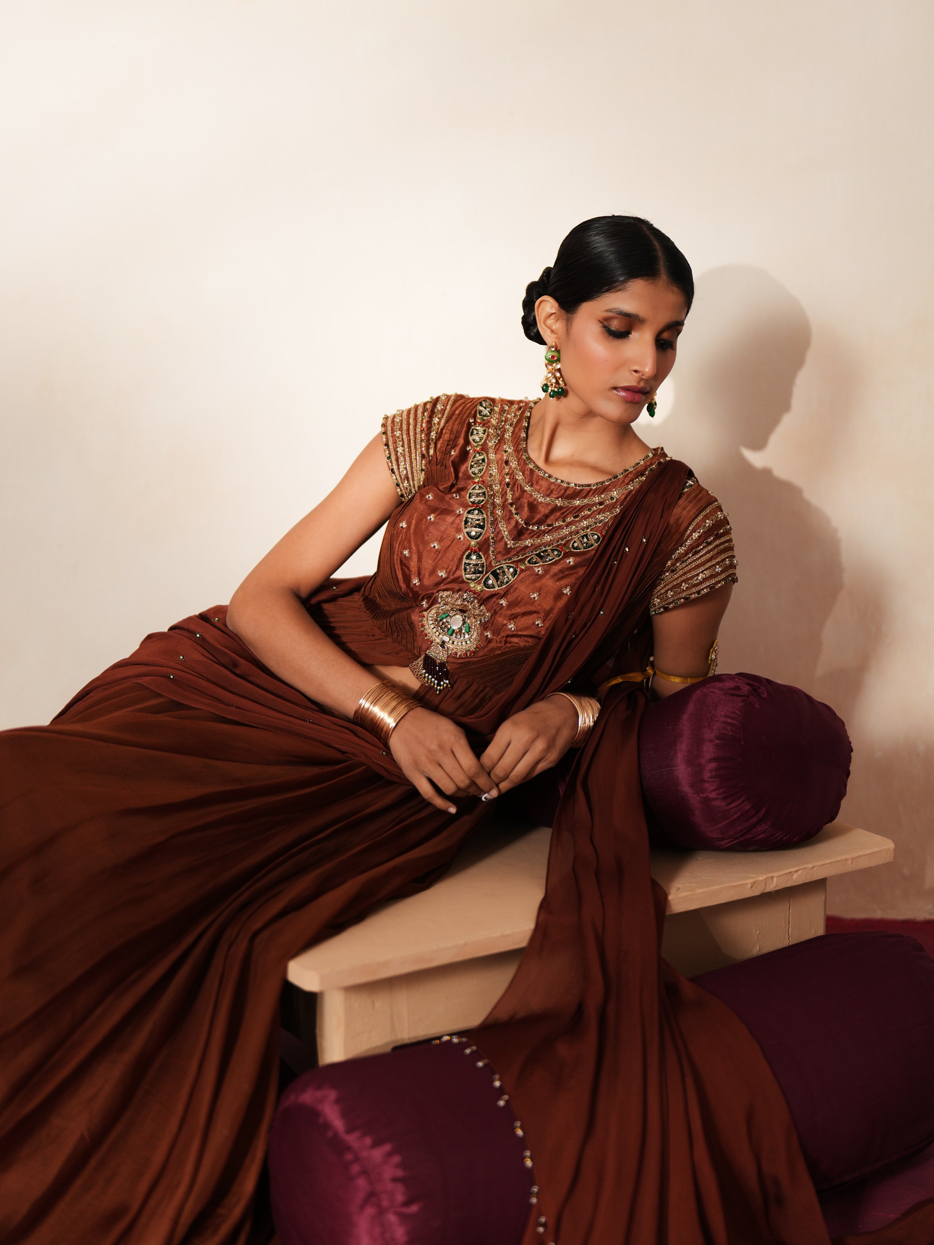 Woman in a mettalic brown traditional outfit standing againsta beige wall on a red carpet.