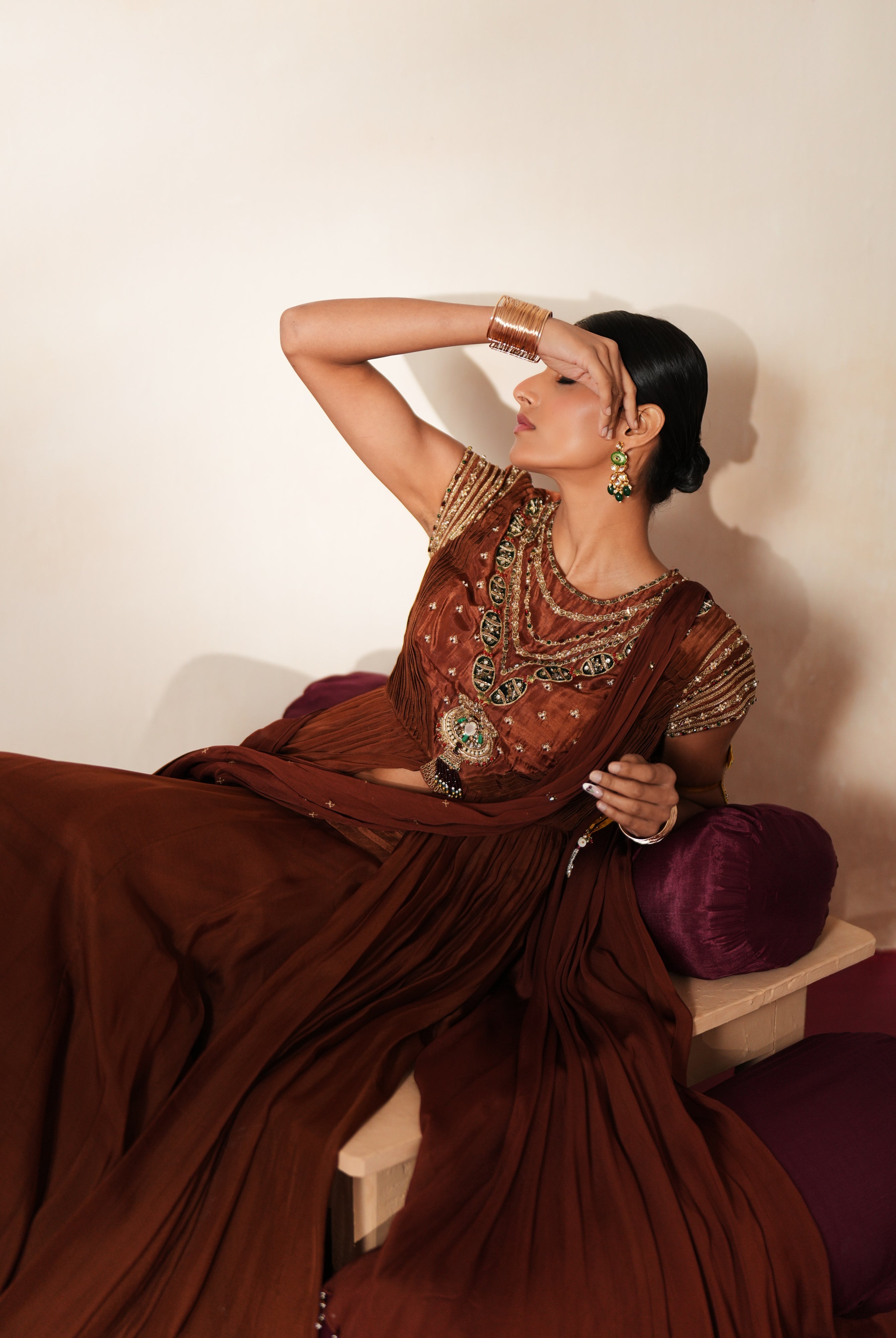 Woman in a mettalic brown traditional outfit standing againsta beige wall on a red carpet.