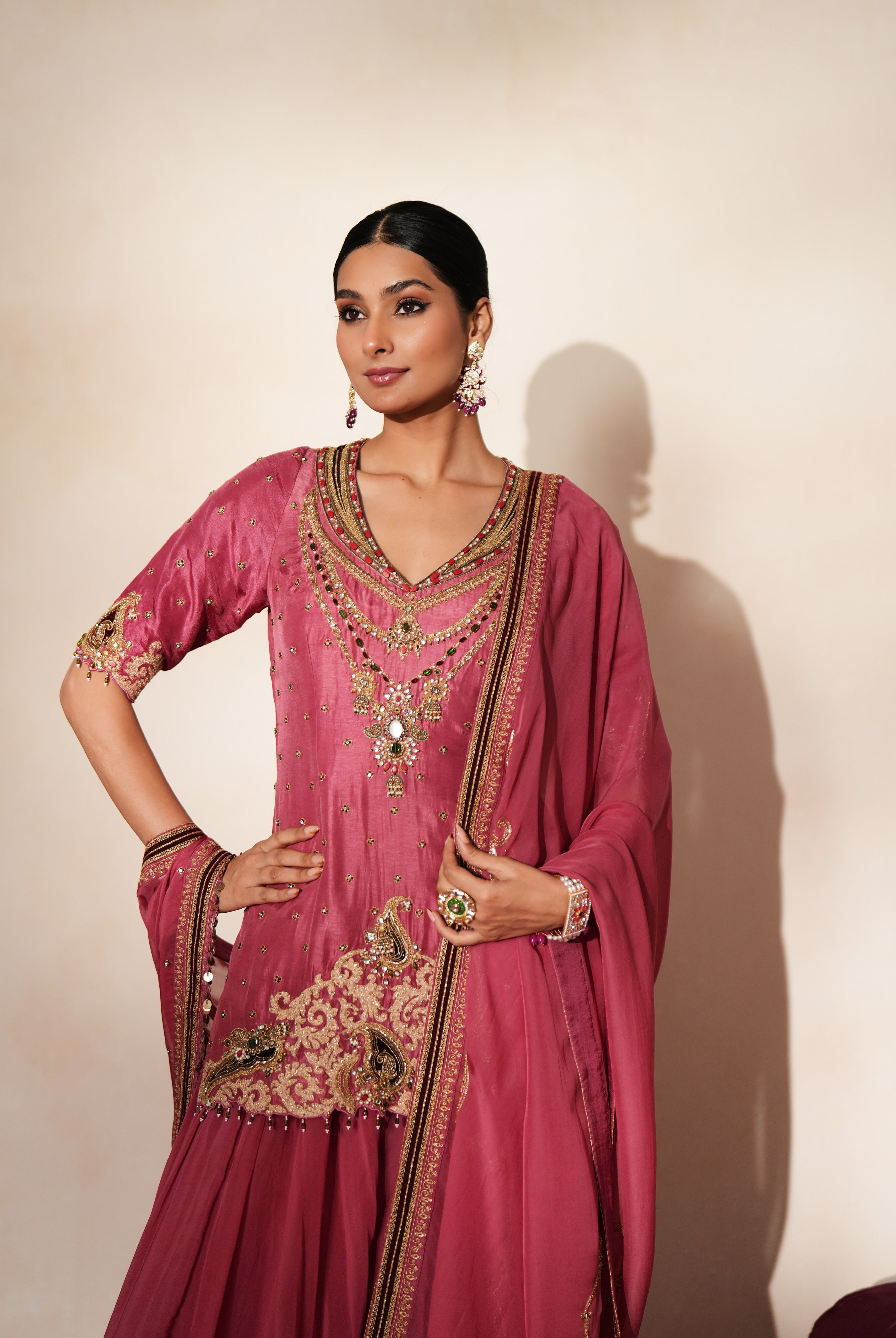 Woman in a mauve pink traditional outfit standing againsta beige wall on a red carpet.