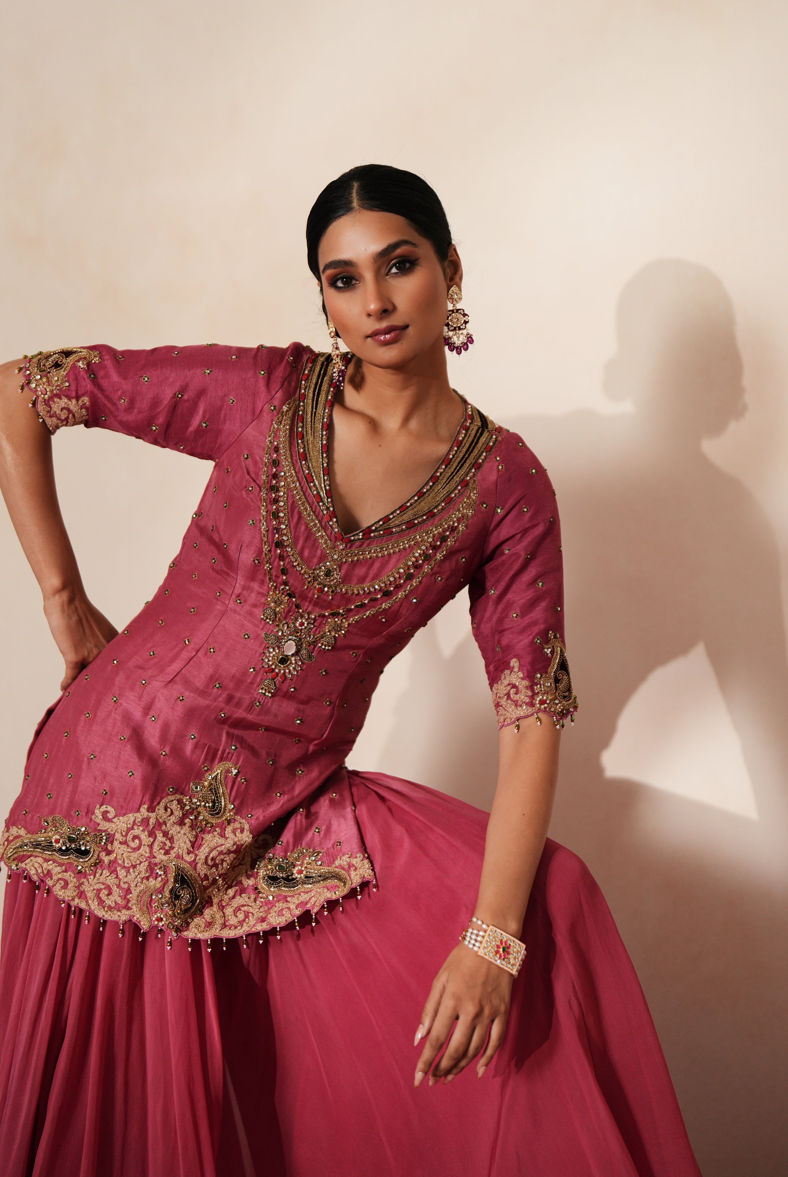 Woman in a mauve pink traditional outfit standing againsta beige wall on a red carpet.
