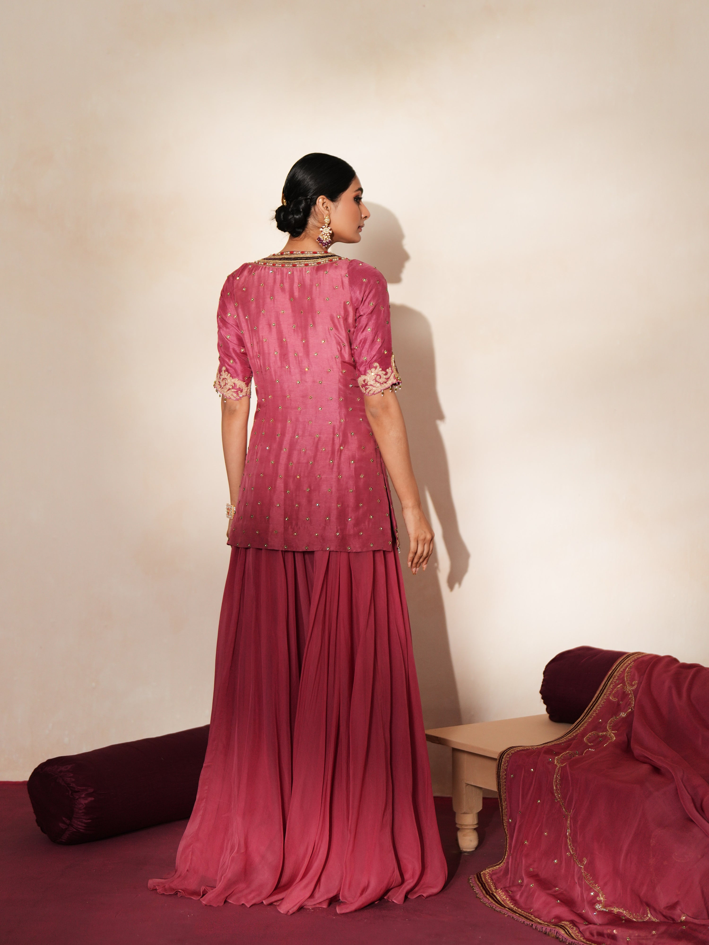 Woman in a mauve pink traditional outfit standing againsta beige wall on a red carpet.