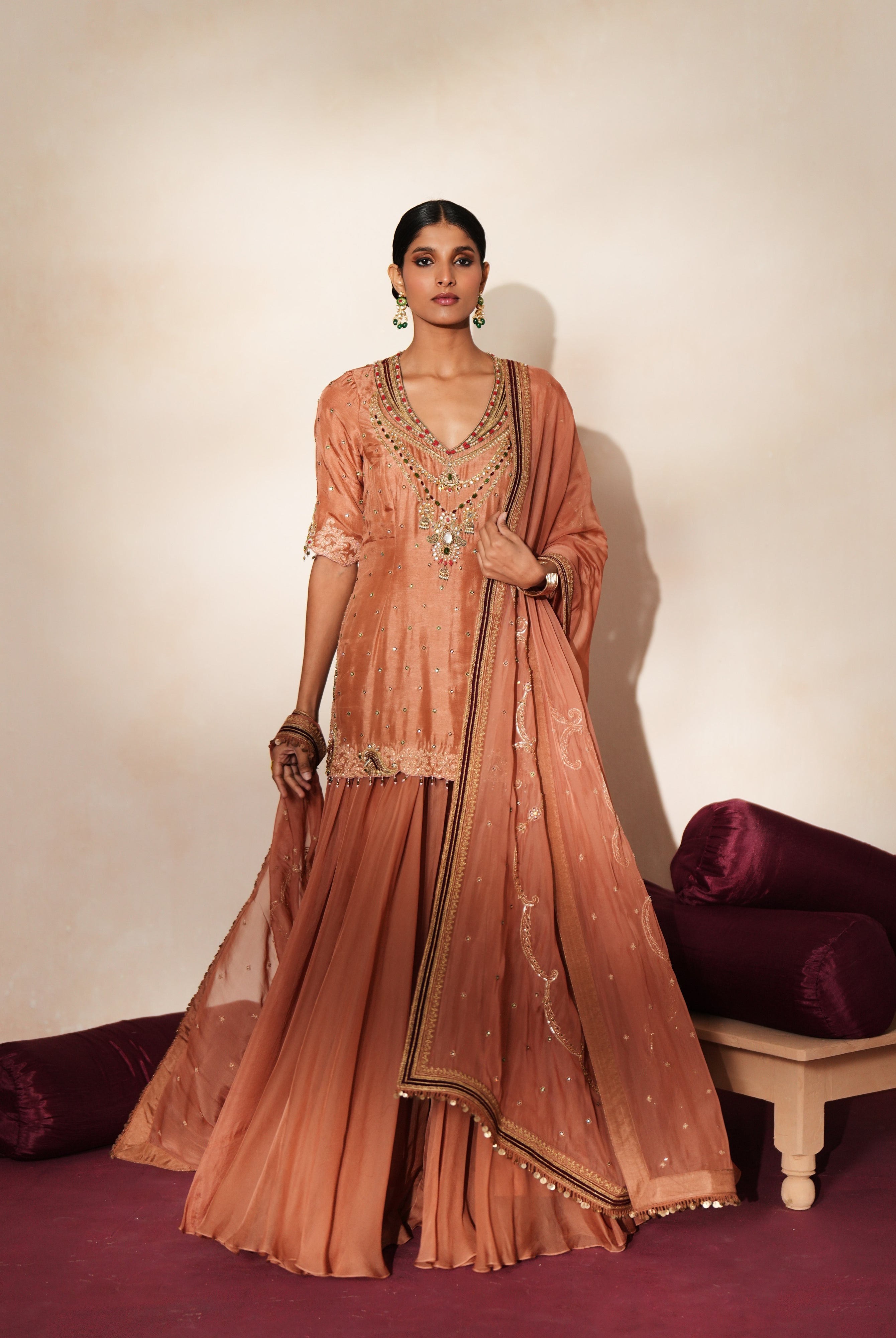 Woman in a metallic mocha traditional outfit standing againsta beige wall on a red carpet.