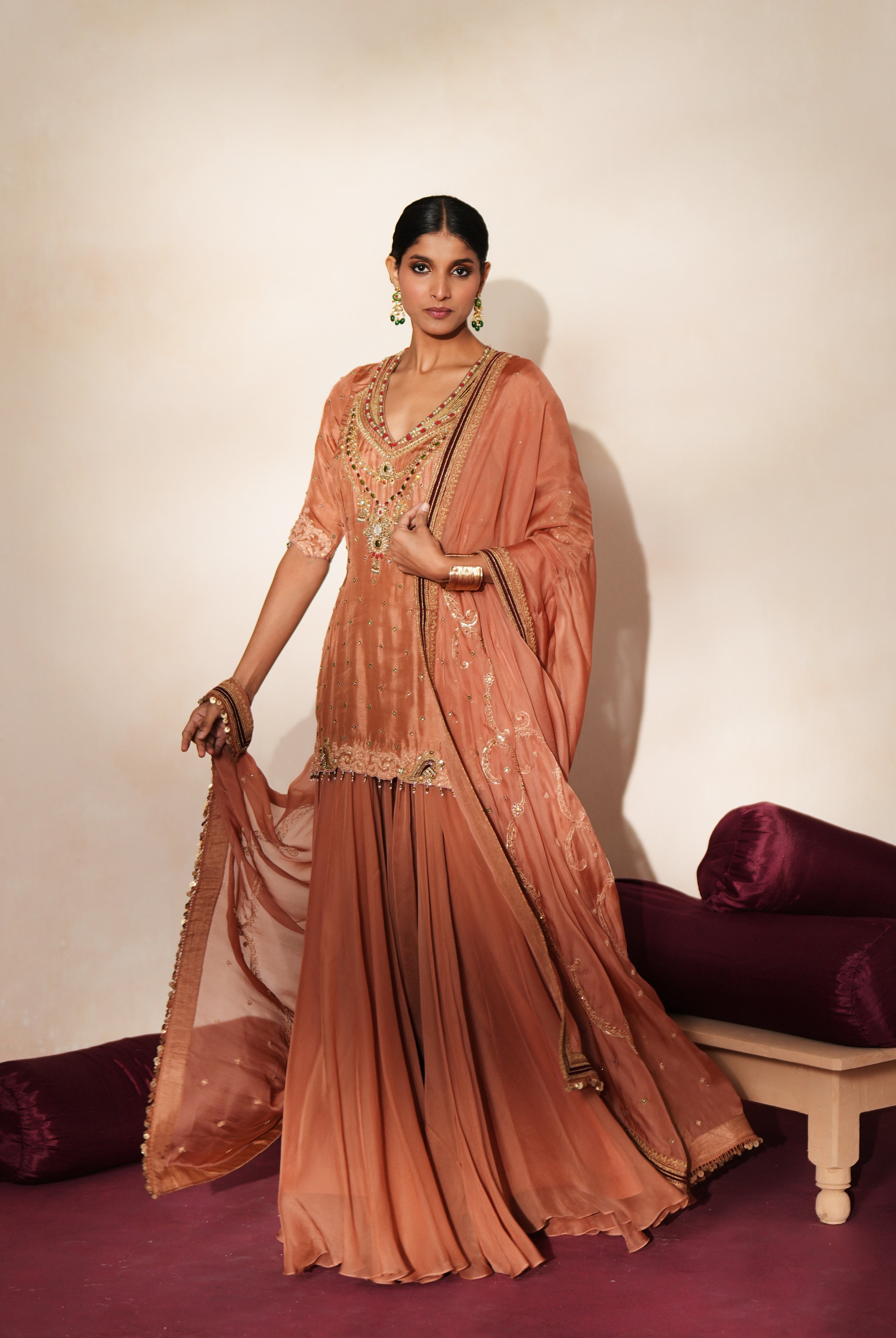 Woman in a metallic mocha traditional outfit standing againsta beige wall on a red carpet.