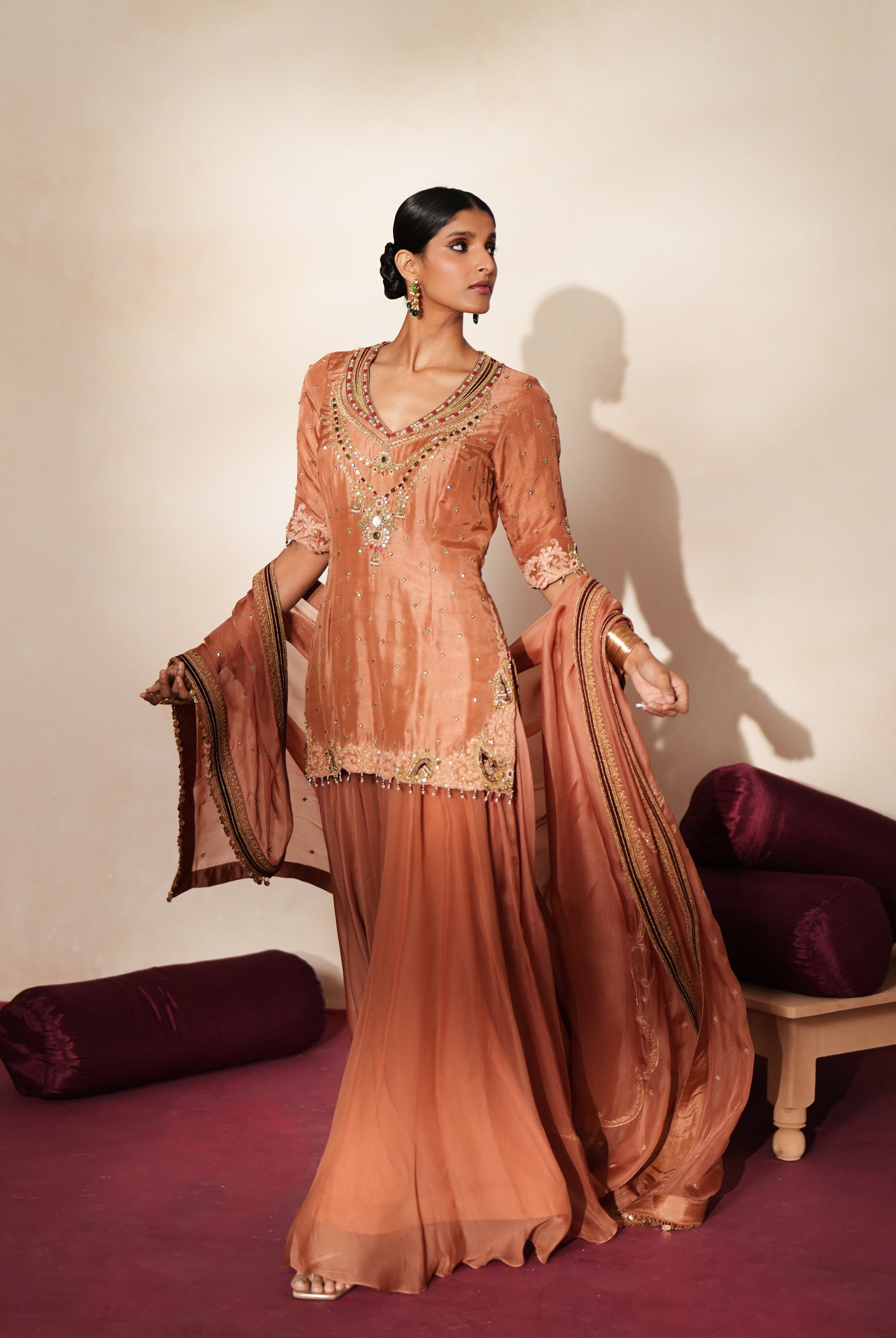 Woman in a metallic mocha traditional outfit standing againsta beige wall on a red carpet.