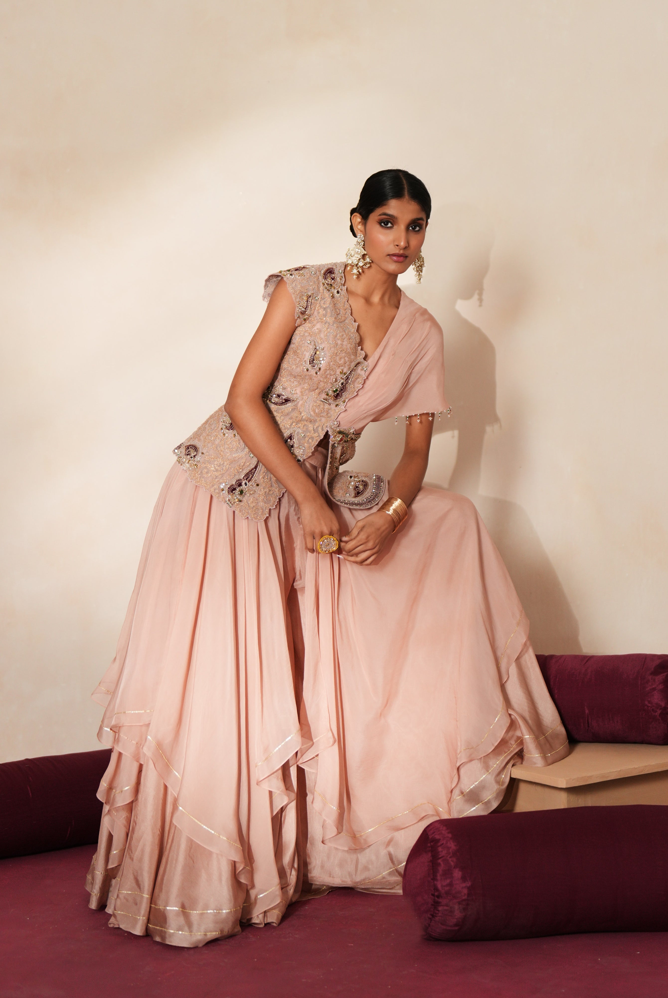 Woman in a soft pink traditional outfit standing againsta beige wall on a red carpet.