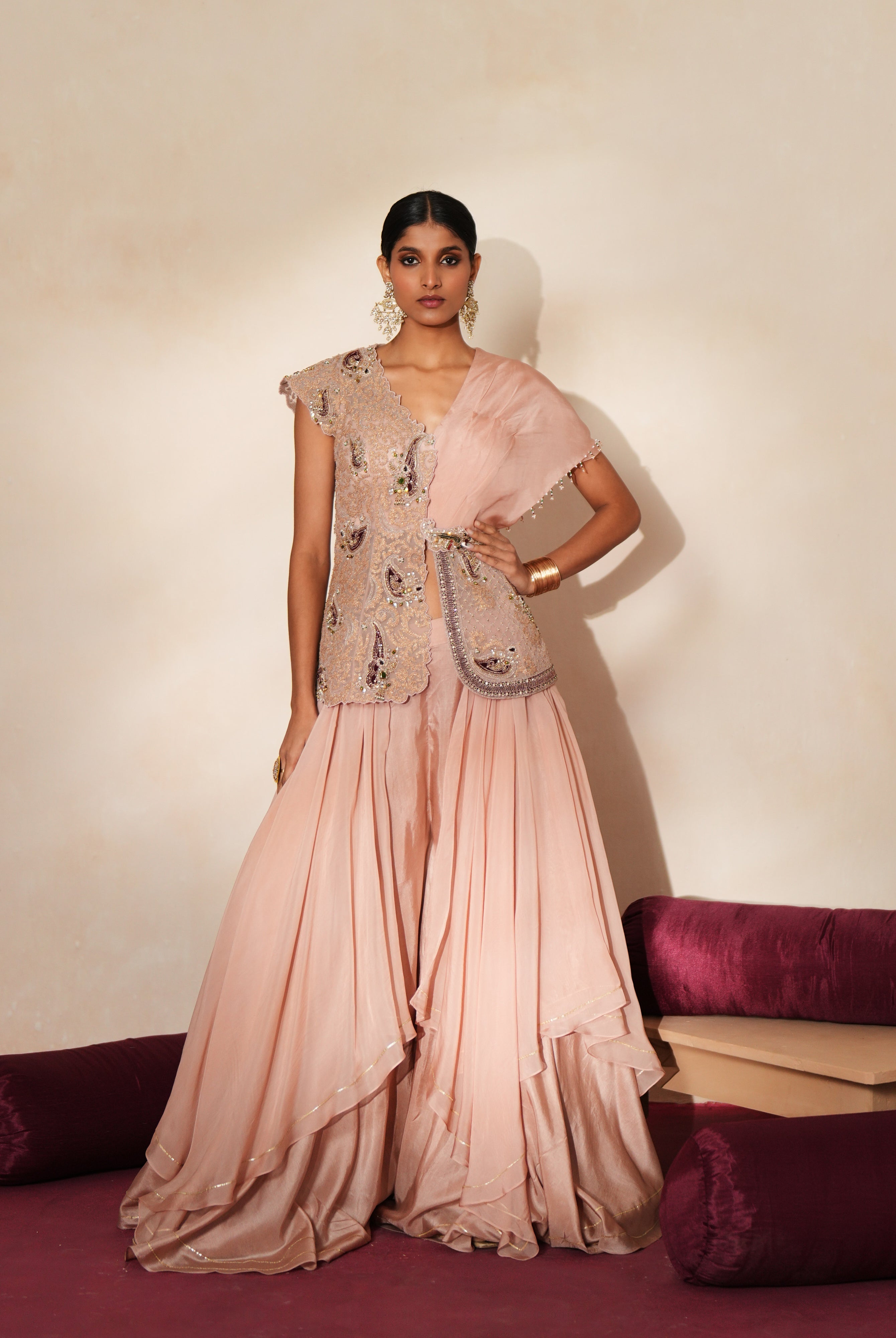 Woman in a soft pink traditional outfit standing againsta beige wall on a red carpet.