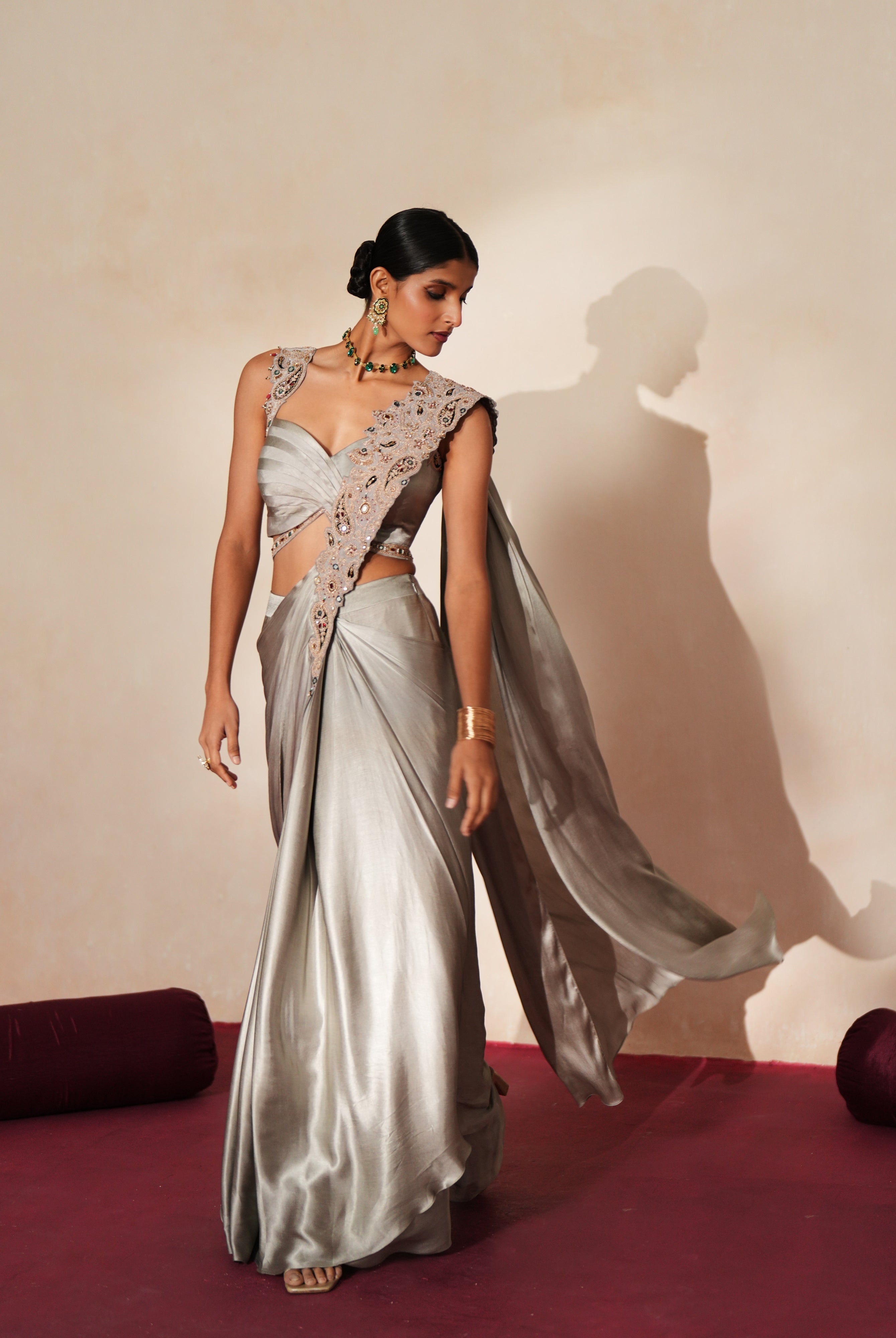 Woman in a mettalic grey traditional outfit standing againsta beige wall on a red carpet.