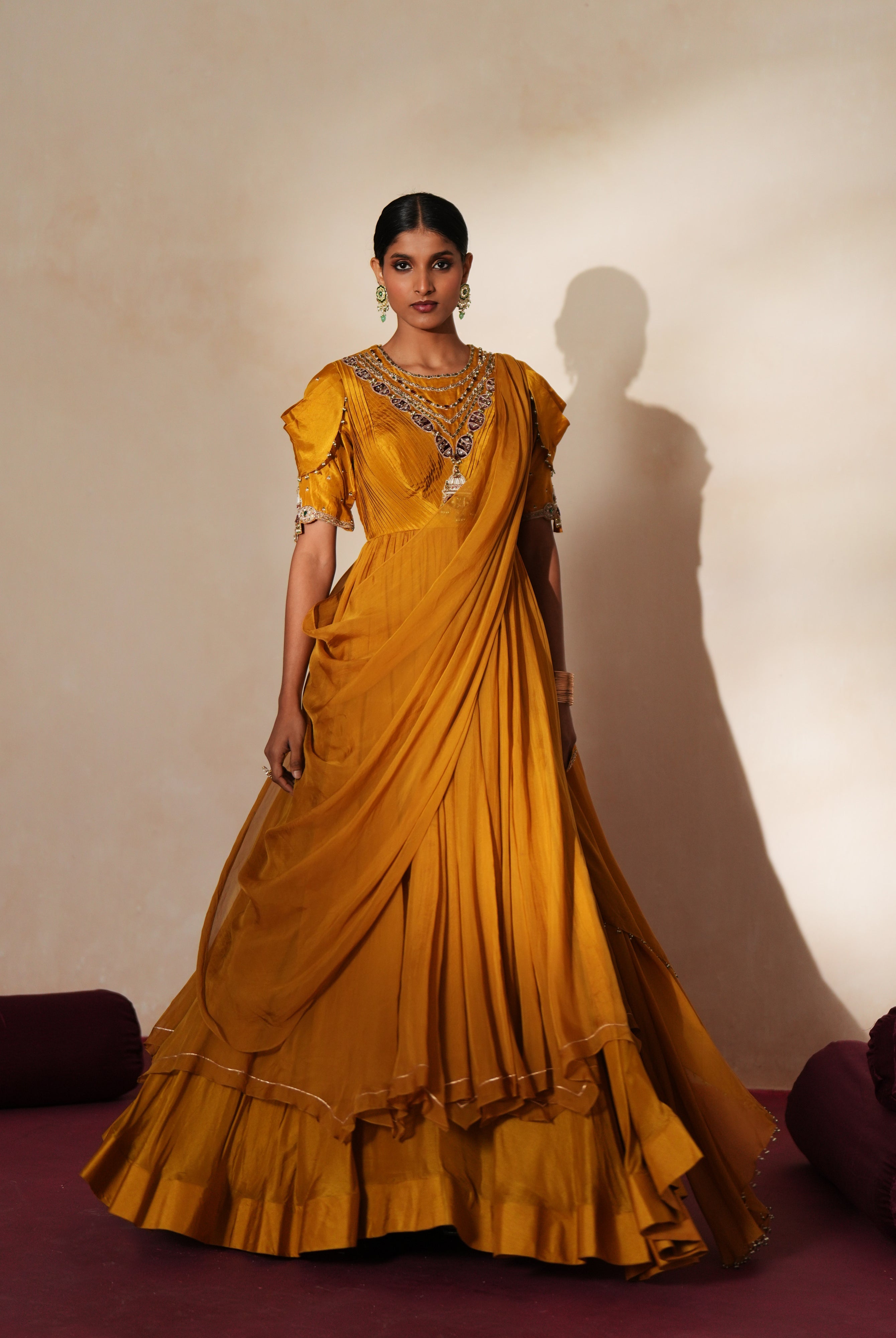 Woman in a golden yellow traditional outfit standing againsta beige wall on a red carpet.