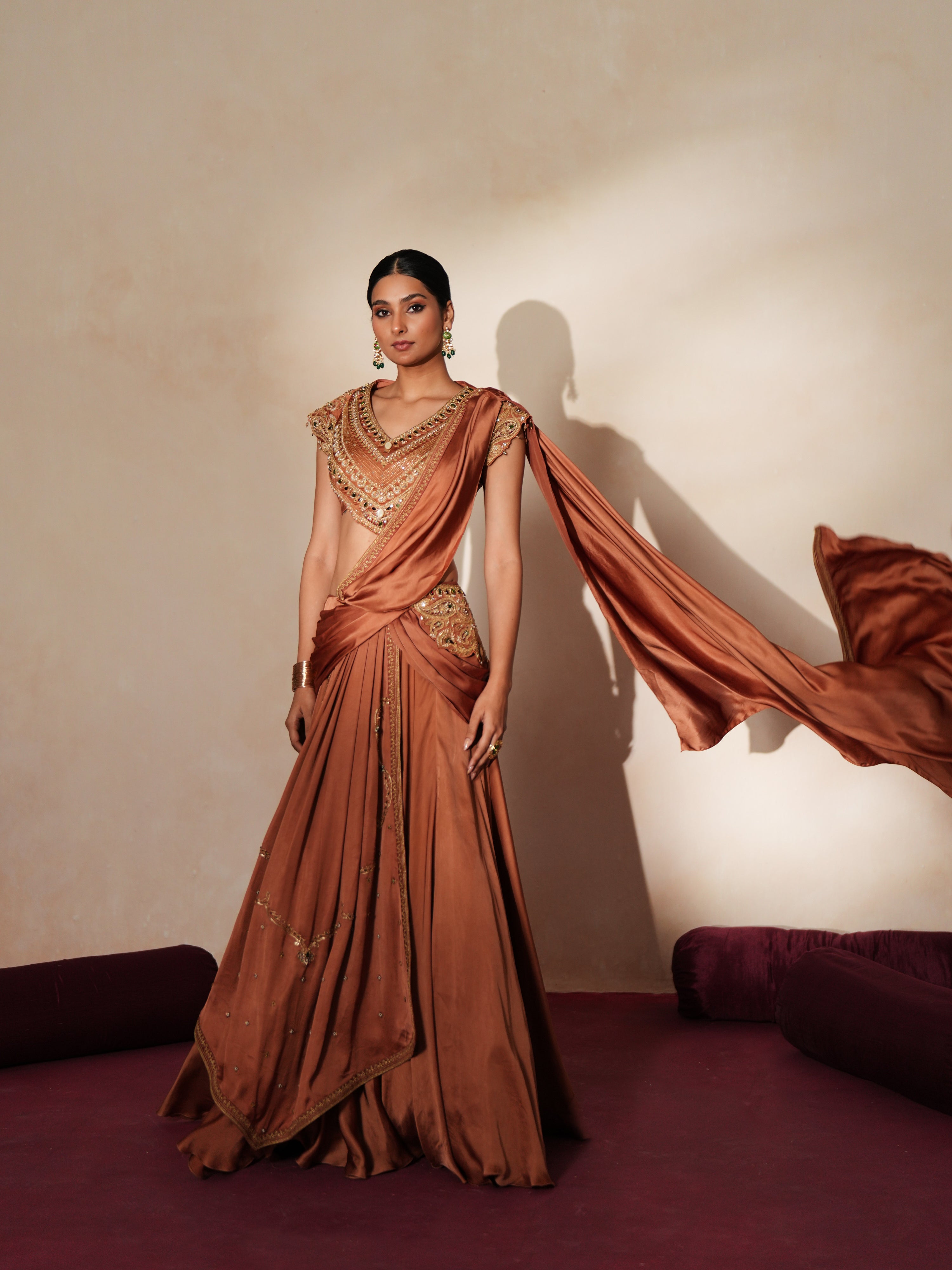 Woman in a metallic mocha traditional outfit standing againsta beige wall on a red carpet.
