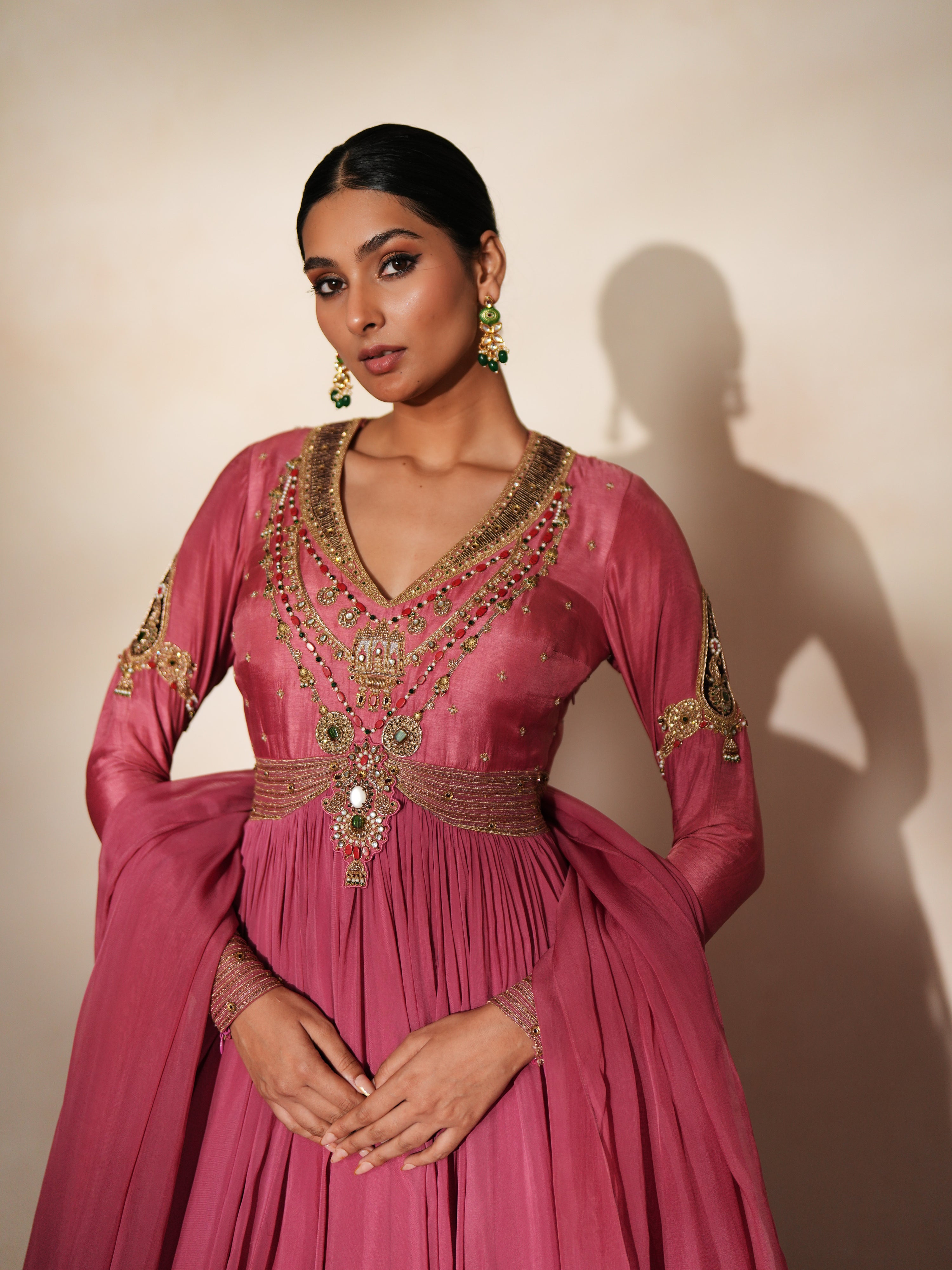 Woman in a mauve pink traditional outfit standing againsta beige wall on a red carpet.