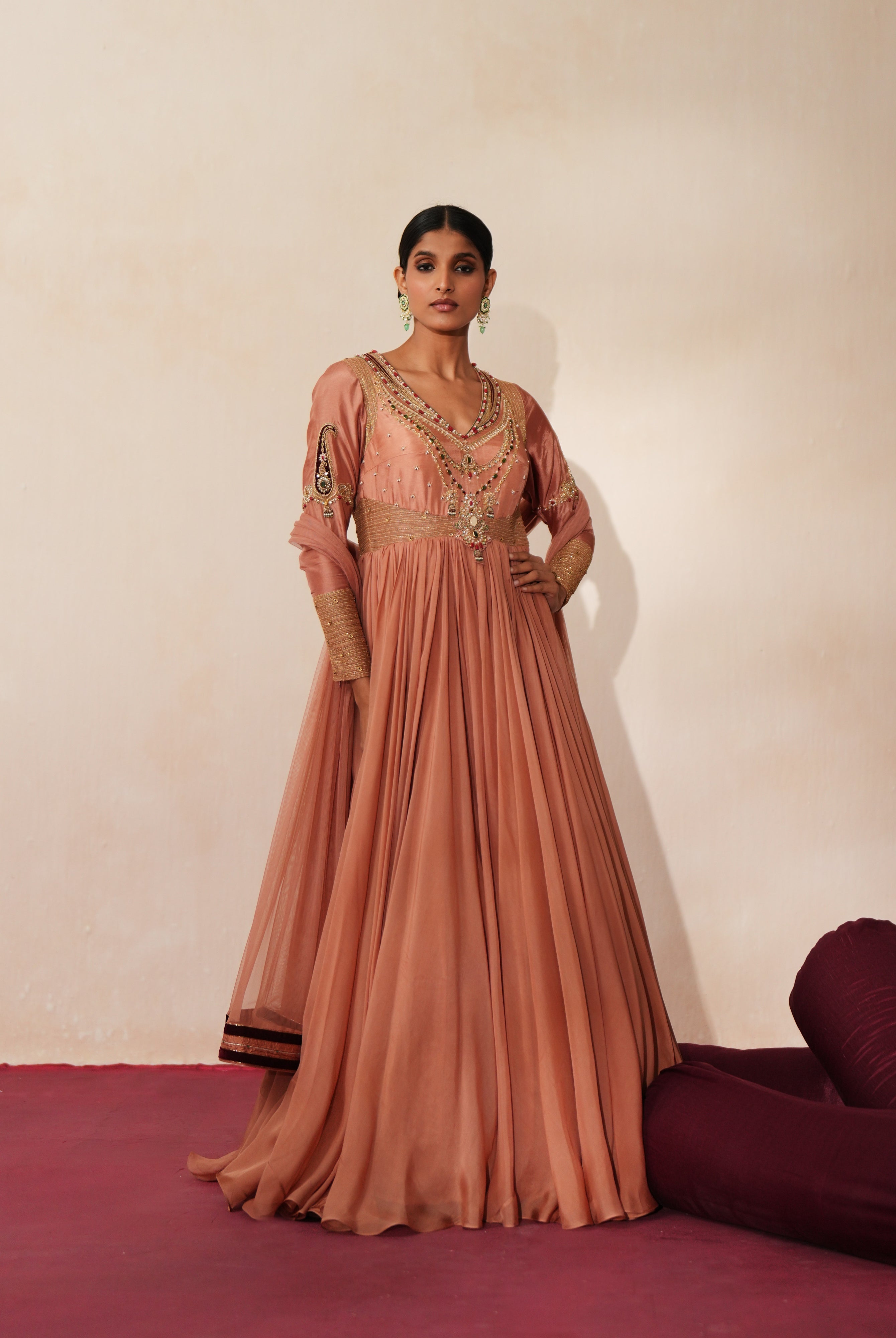 Woman in a metallic mocha traditional outfit standing againsta beige wall on a red carpet.