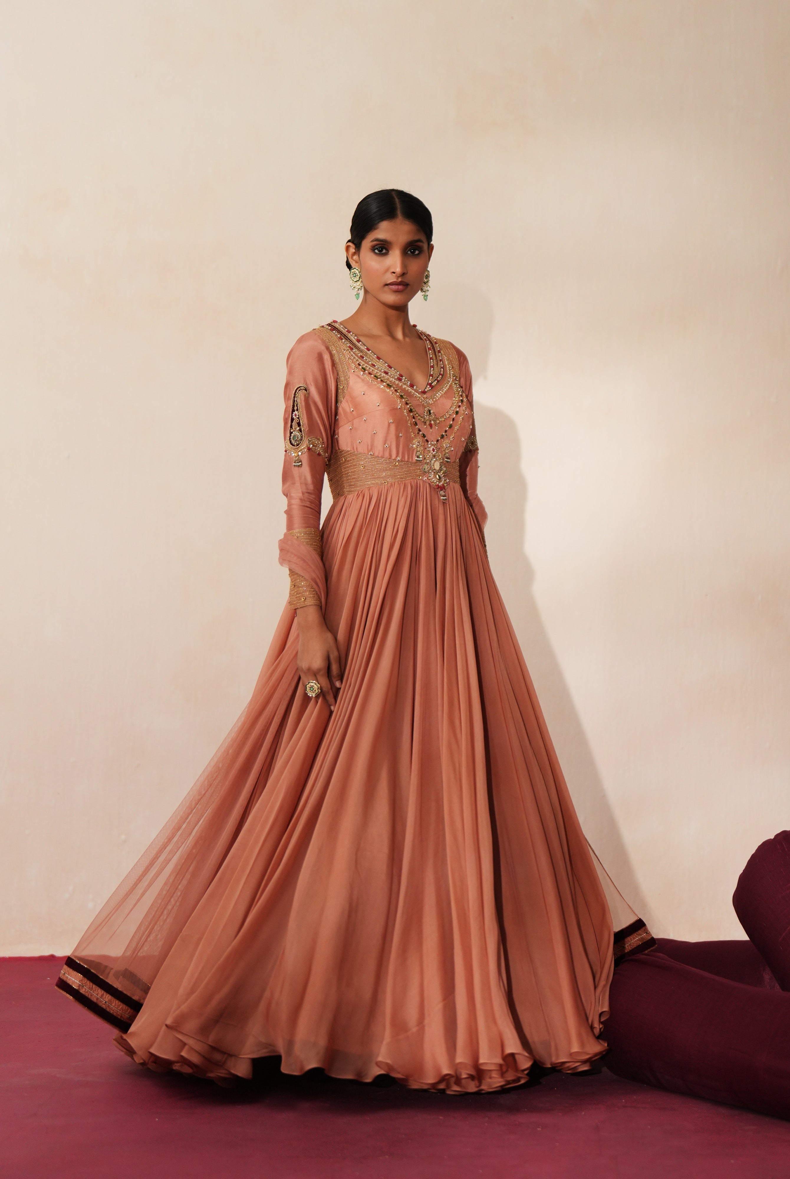 Woman in a metallic mocha traditional outfit standing againsta beige wall on a red carpet.