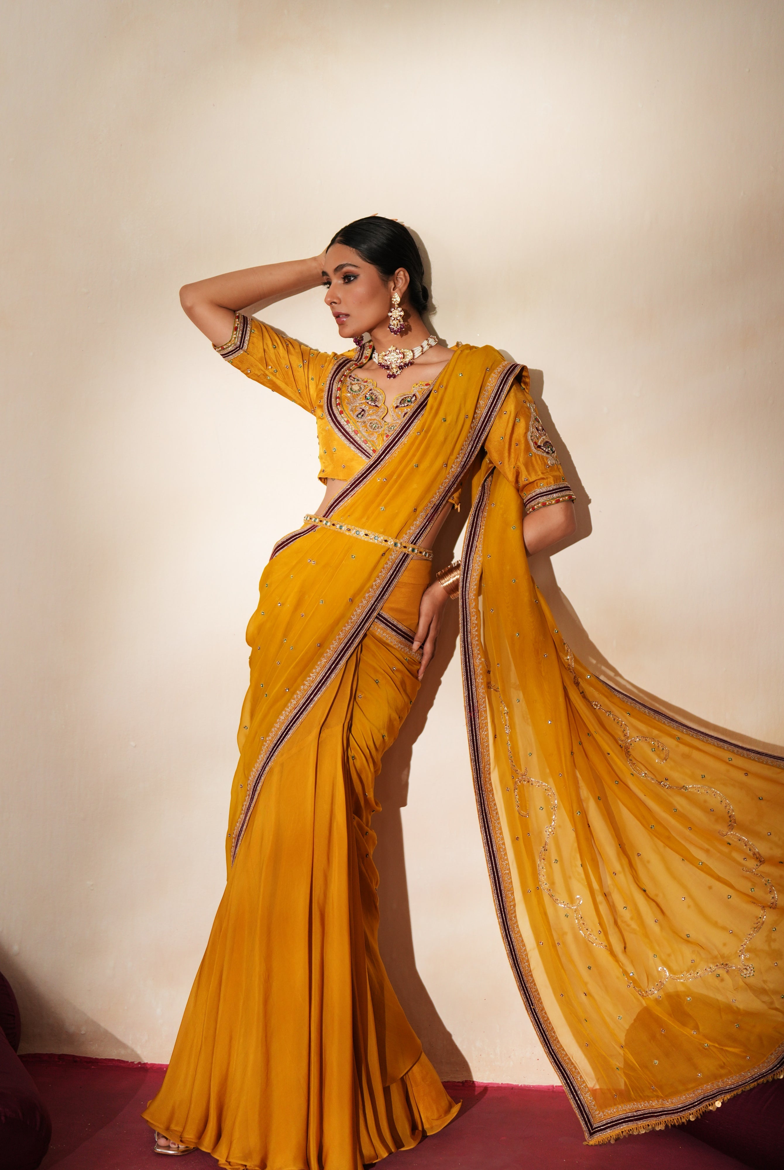 Woman in a golden yellow traditional outfit standing againsta beige wall on a red carpet.