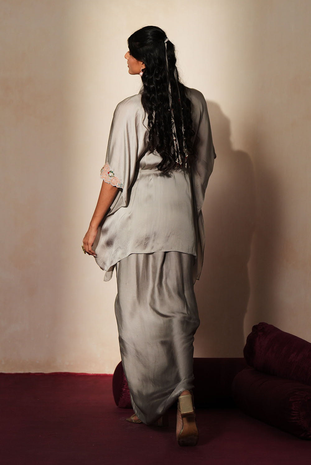 Woman in a mettalic grey traditional outfit standing againsta beige wall on a red carpet.