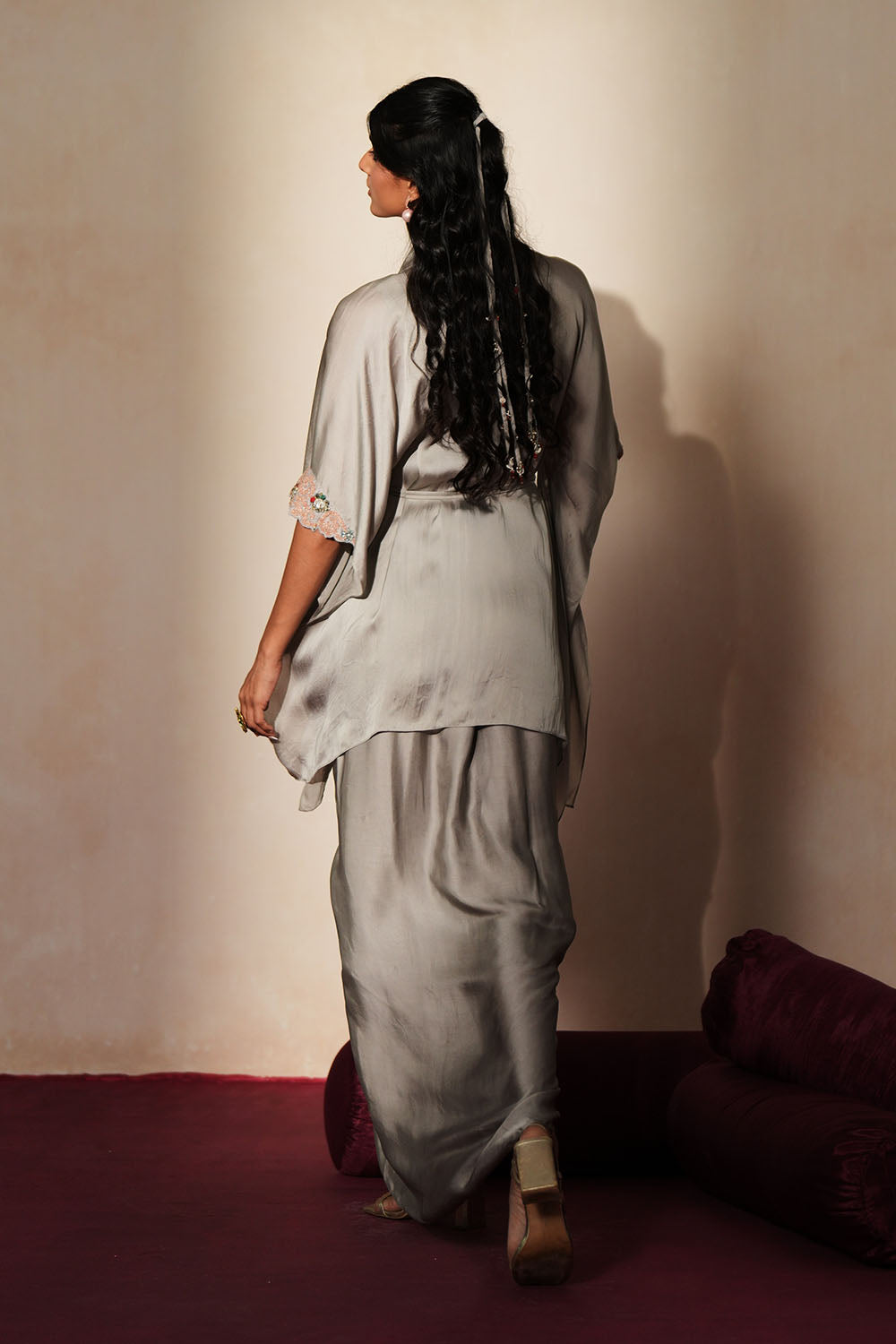 Woman in a mettalic grey traditional outfit standing againsta beige wall on a red carpet.