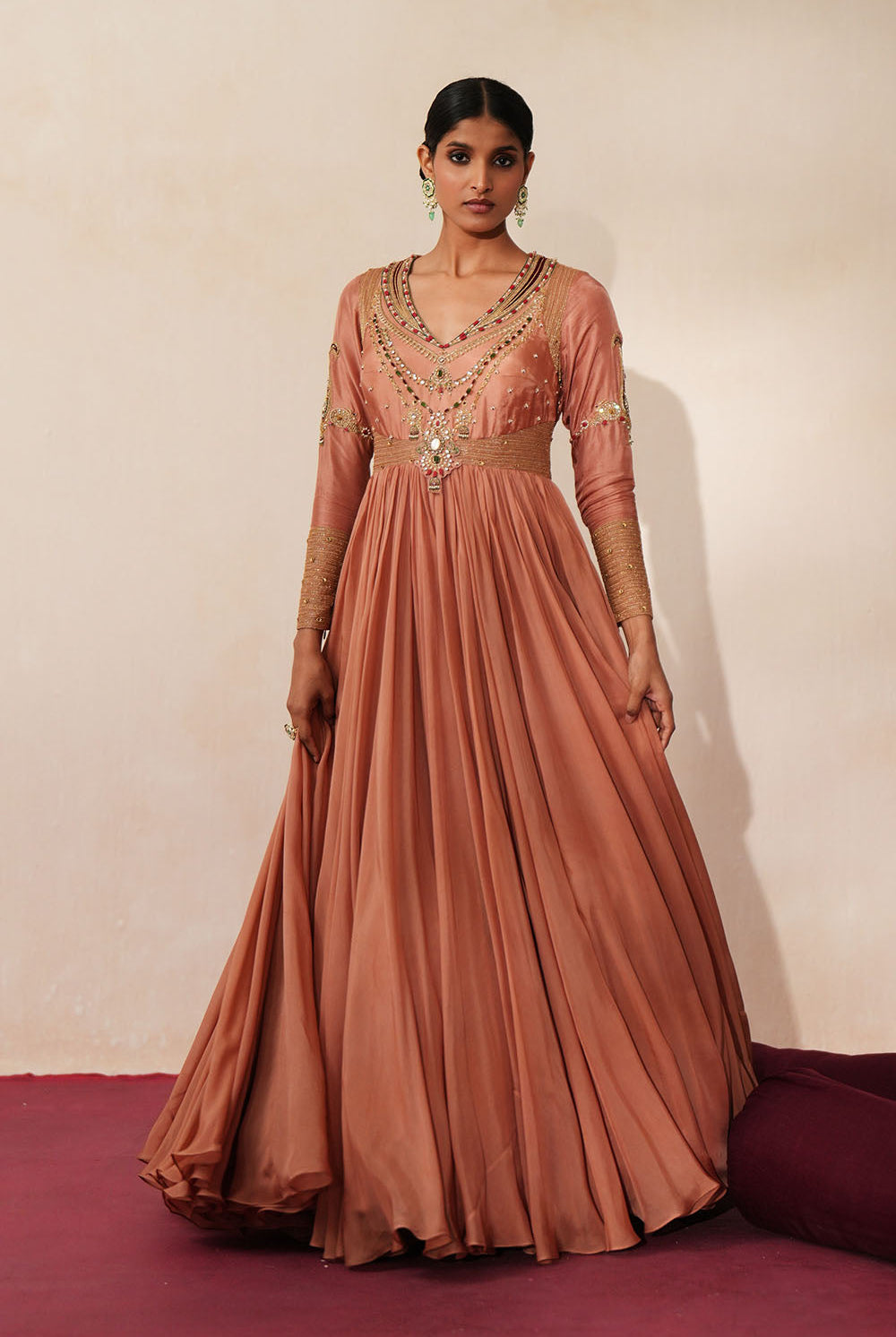 Woman in a metallic mocha traditional outfit standing againsta beige wall on a red carpet.