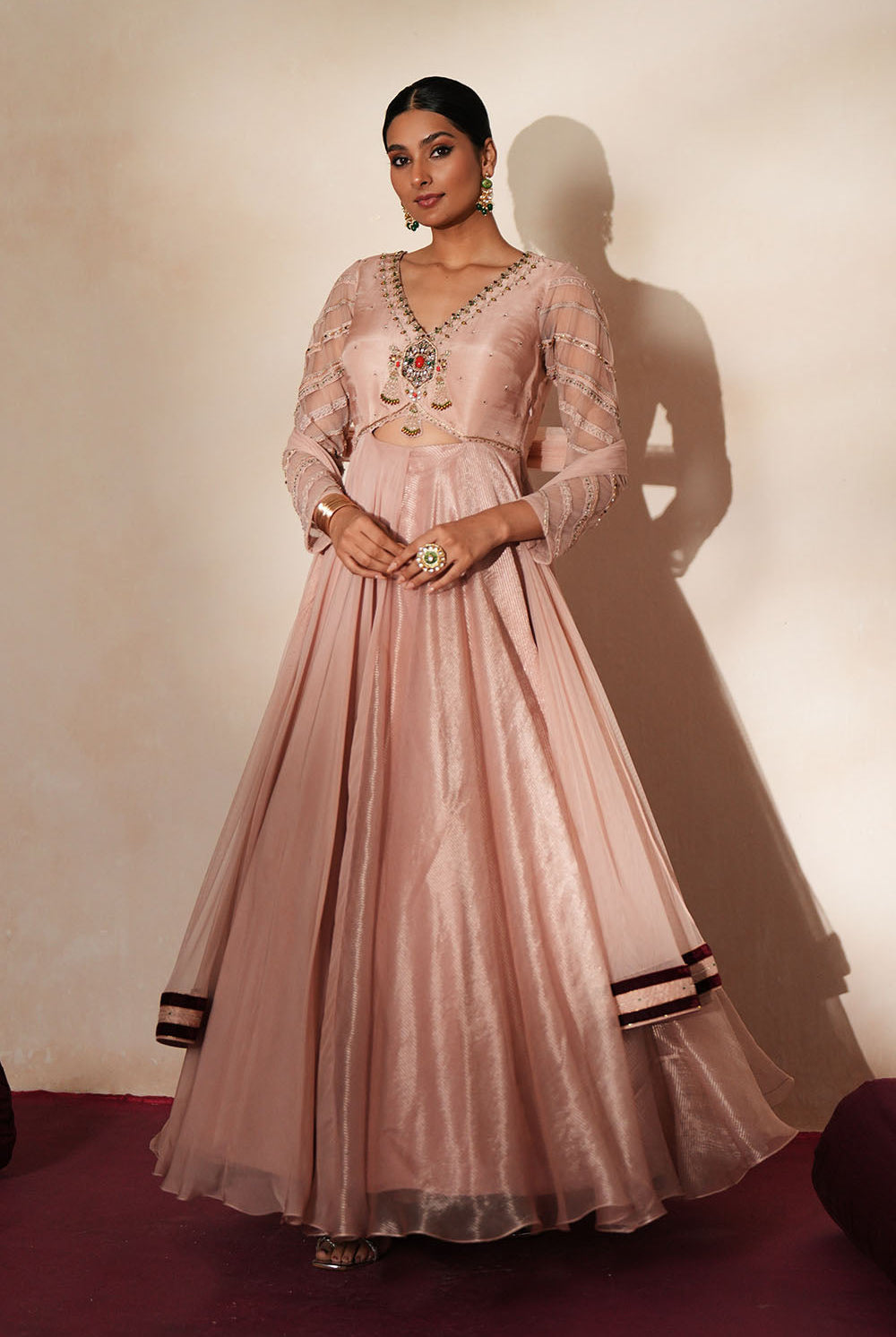 Woman in a soft pink traditional outfit standing againsta beige wall on a red carpet.