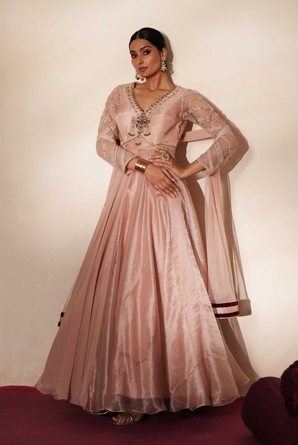 Woman in a soft pink traditional outfit standing againsta beige wall on a red carpet.