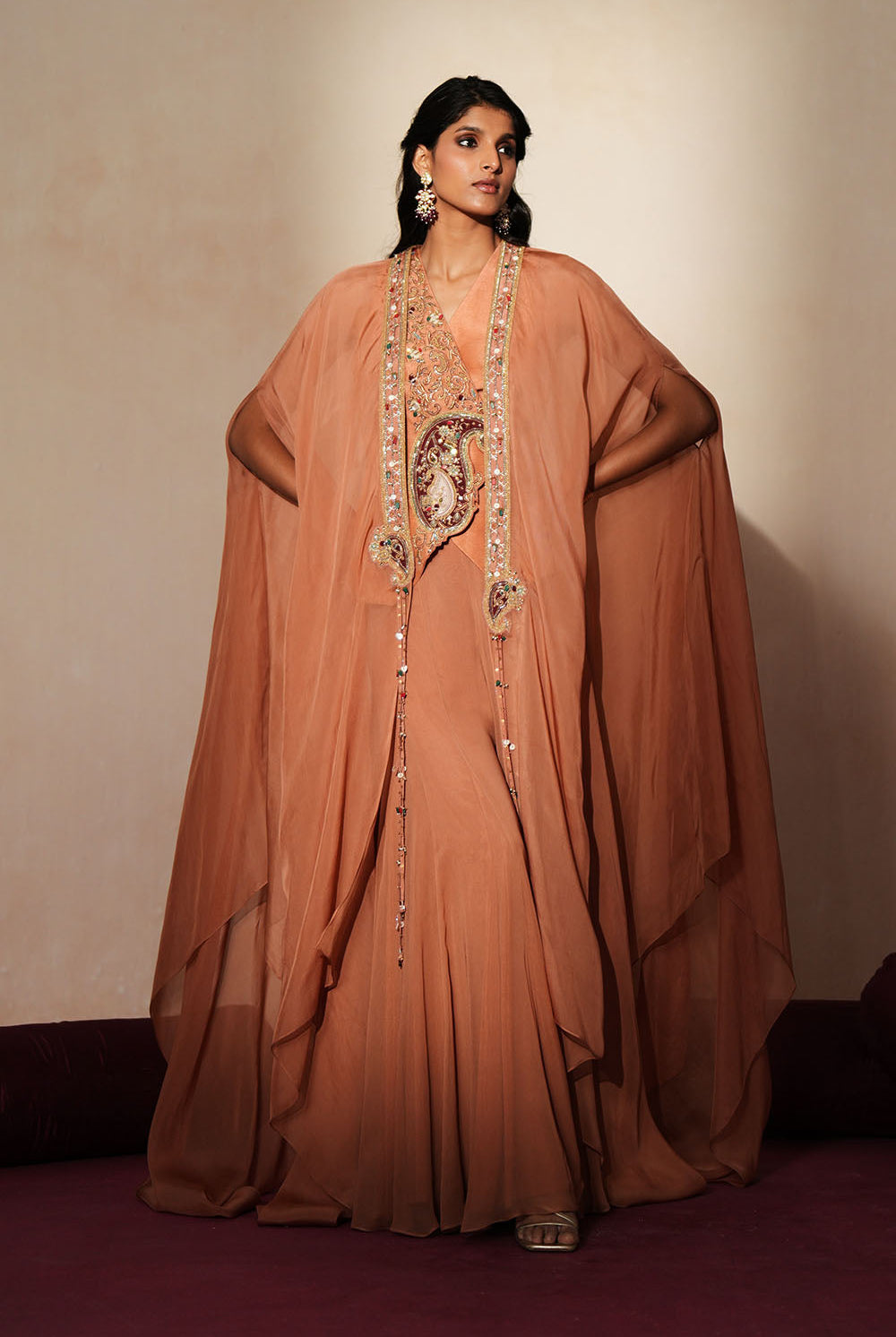 Woman in a metallic mocha traditional outfit standing againsta beige wall on a red carpet.