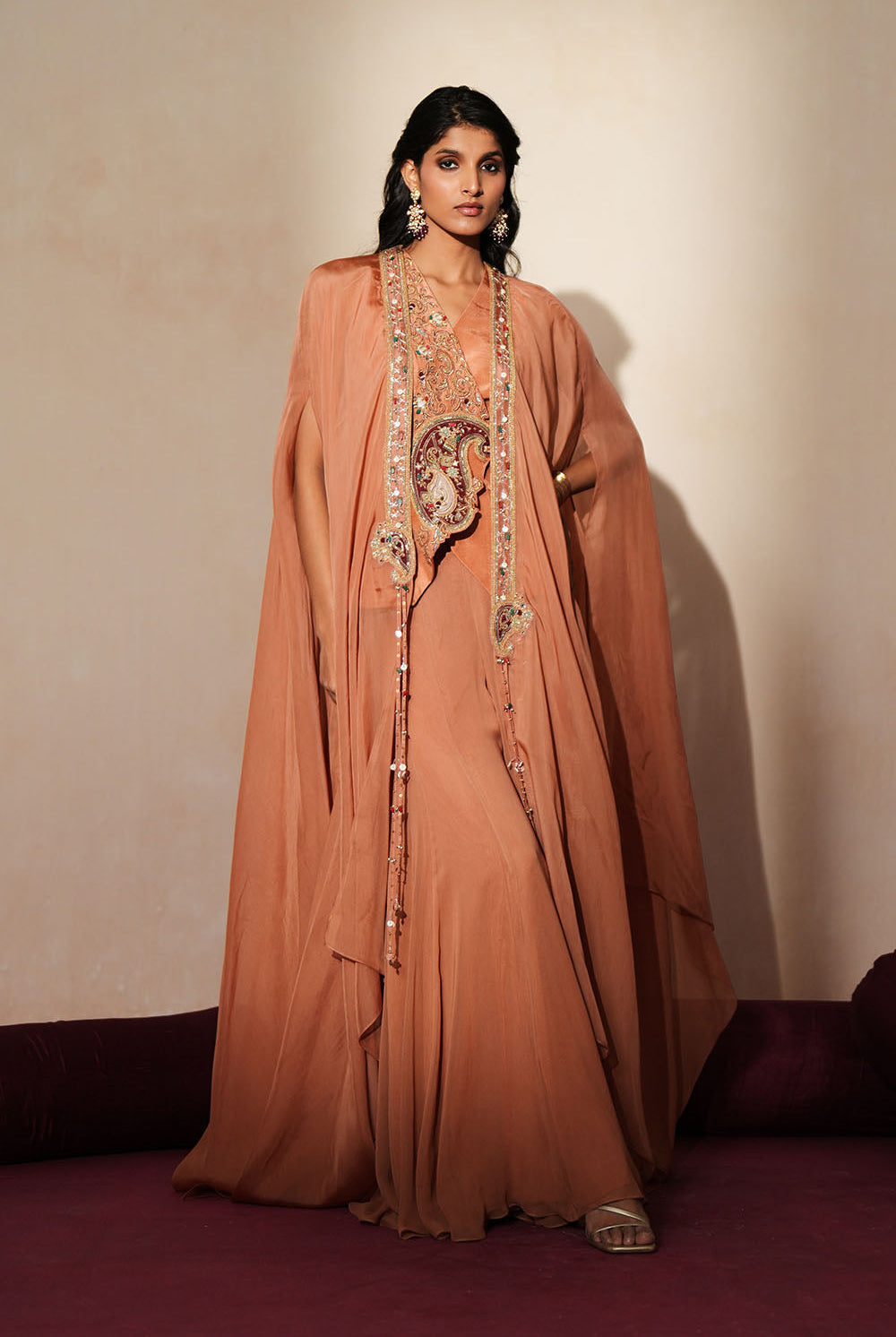 Woman in a metallic mocha traditional outfit standing againsta beige wall on a red carpet.