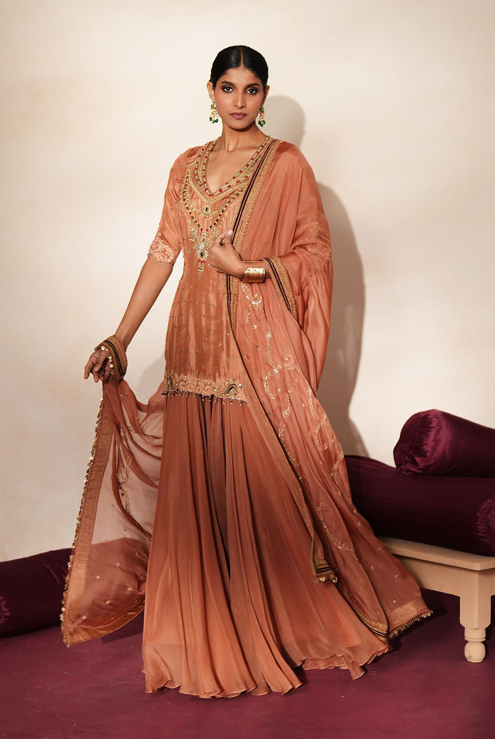 Woman in a metallic mocha traditional outfit standing againsta beige wall on a red carpet.