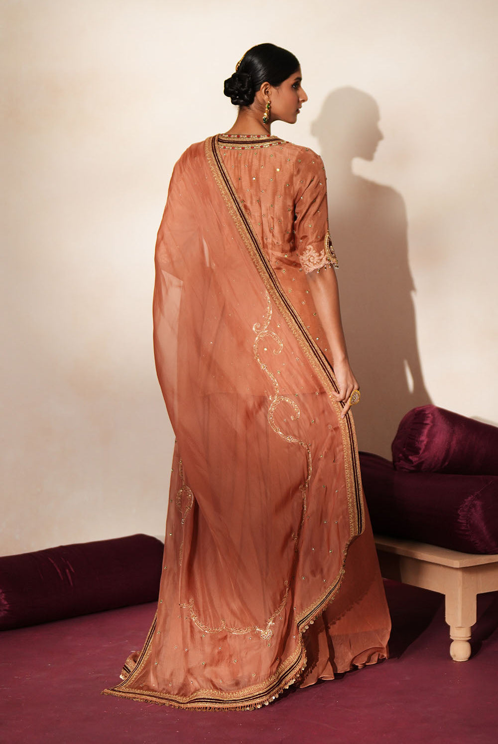Woman in a metallic mocha traditional outfit standing againsta beige wall on a red carpet.