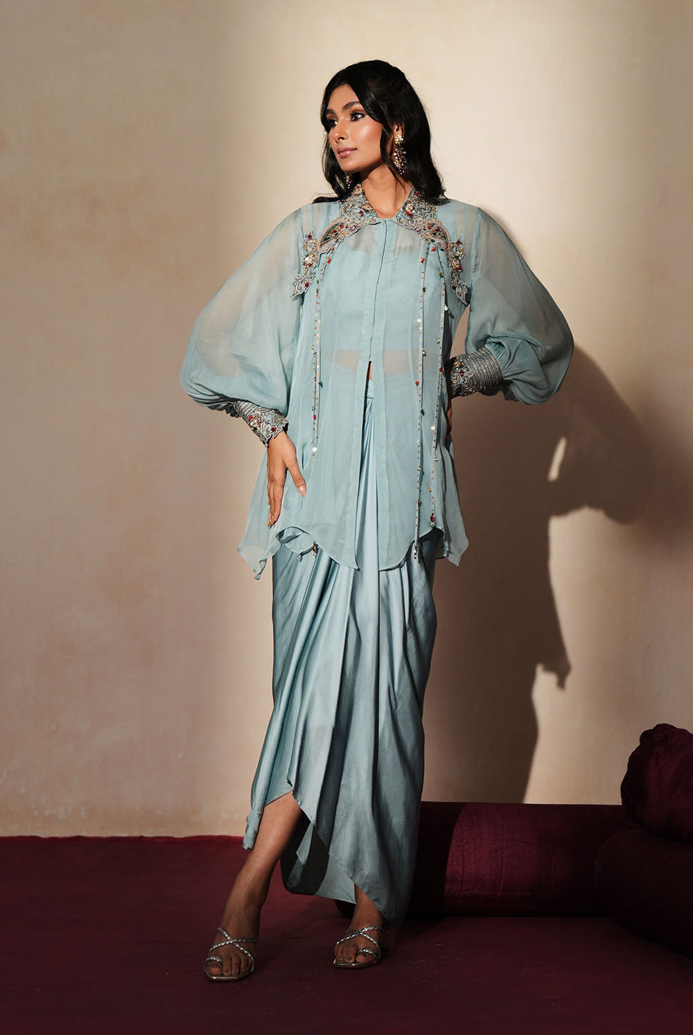 Woman in a pastel blue traditional outfit standing againsta beige wall on a red carpet.