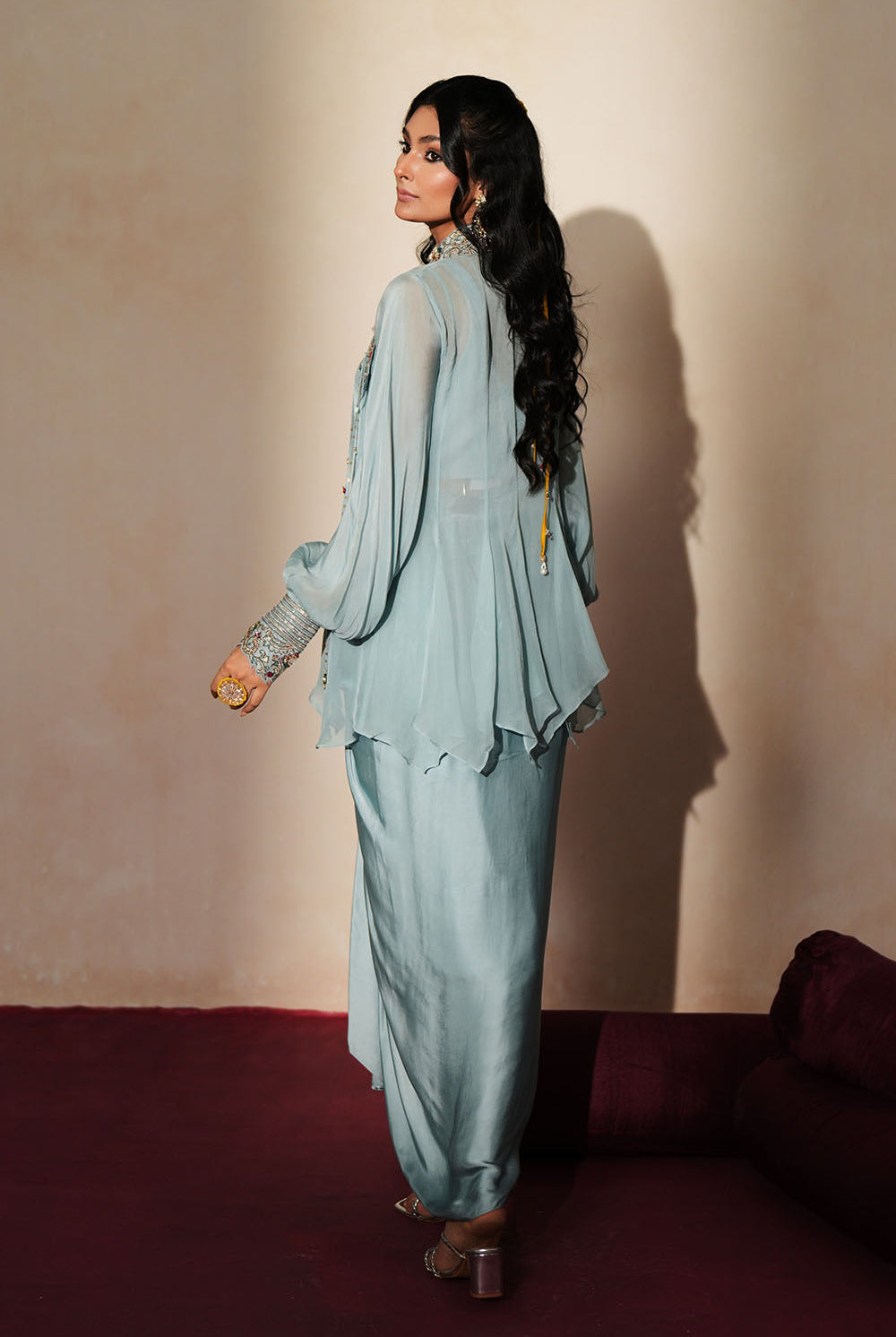 Woman in a pastel blue traditional outfit standing againsta beige wall on a red carpet.