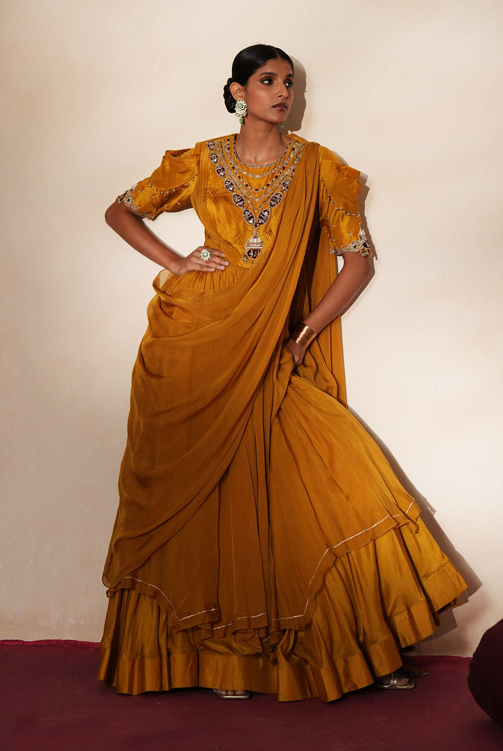 Woman in a golden yellow traditional outfit standing againsta beige wall on a red carpet.