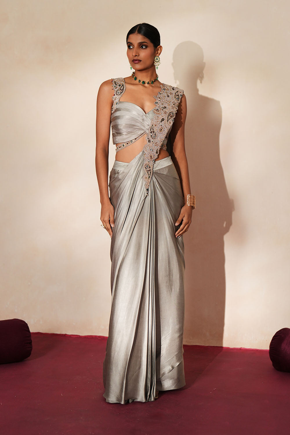 Woman in a mettalic grey traditional outfit standing againsta beige wall on a red carpet.