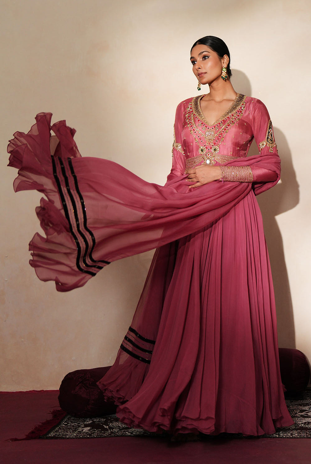 Woman in a mauve pink traditional outfit standing againsta beige wall on a red carpet.