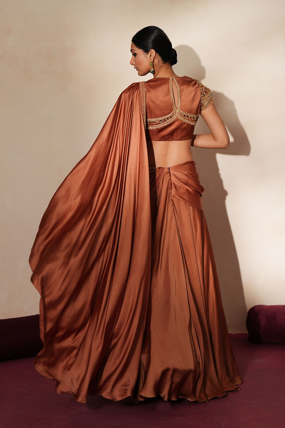 Woman in a metallic mocha traditional outfit standing againsta beige wall on a red carpet.